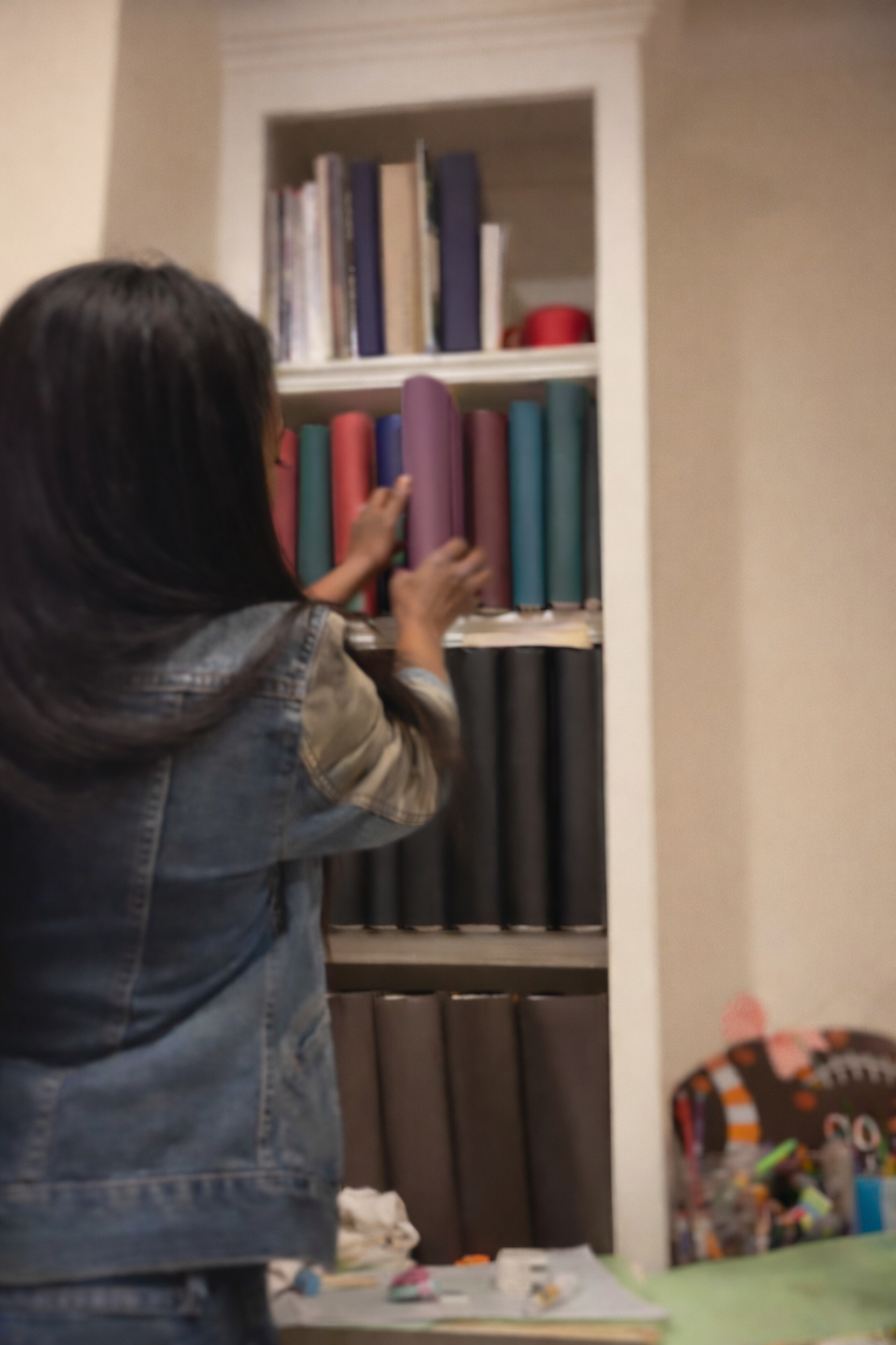 A woman with dark hair, wearing a denim jacket, is reaching for colorful binders on a bookshelf in a room.