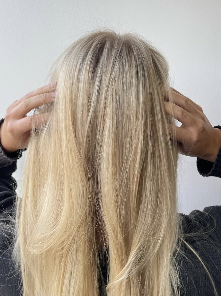 Person with long blonde hair holding their head with hands, facing a white wall.