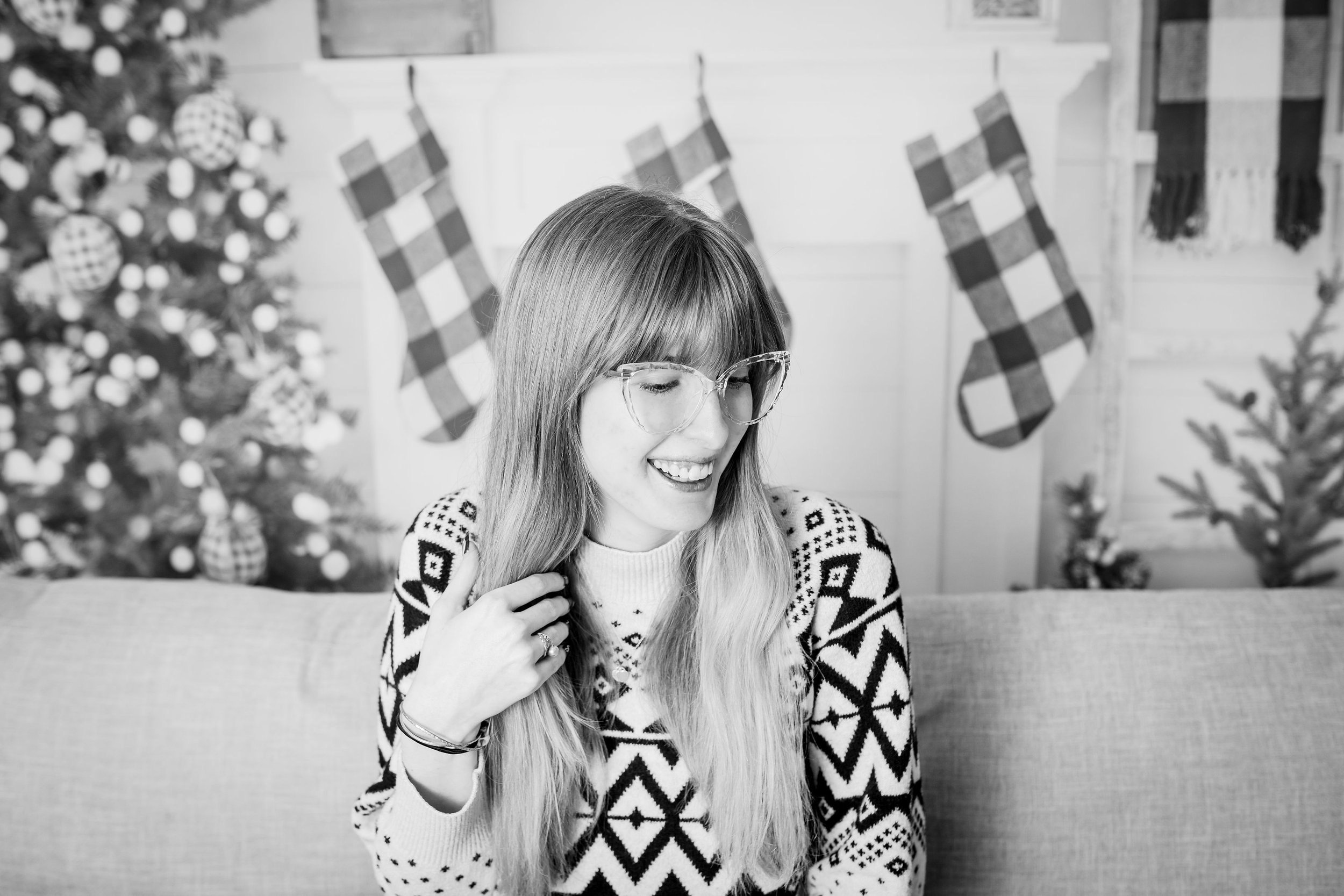 A woman with long hair and glasses sitting on a sofa, smiling, in front of a decorated Christmas scene with stockings hanging on a mantle and a Christmas tree.