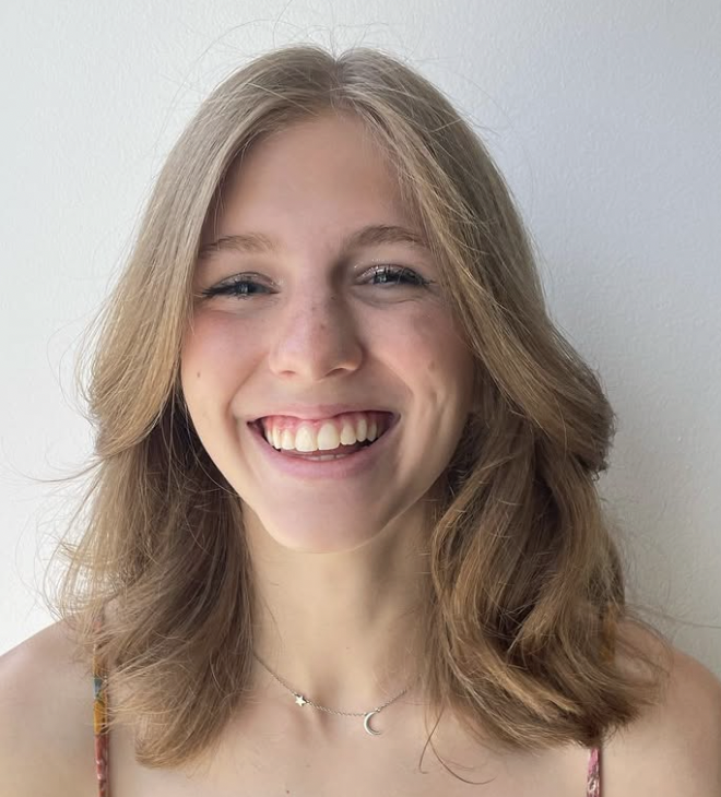 Close-up of a young woman with shoulder-length light brown hair, smiling with visible teeth, wearing a delicate necklace with crescent moon and star charms, standing against a plain white wall.