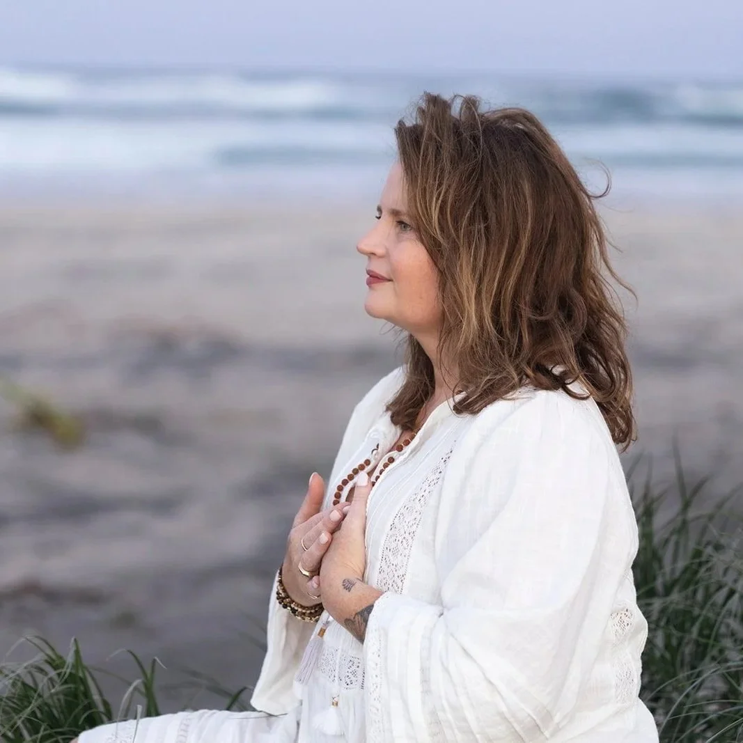 A woman with shoulder-length brown hair sits on the beach with her eyes closed, hands pressed together over her chest, wearing a white dress and a beaded bracelet, with the ocean in the background.