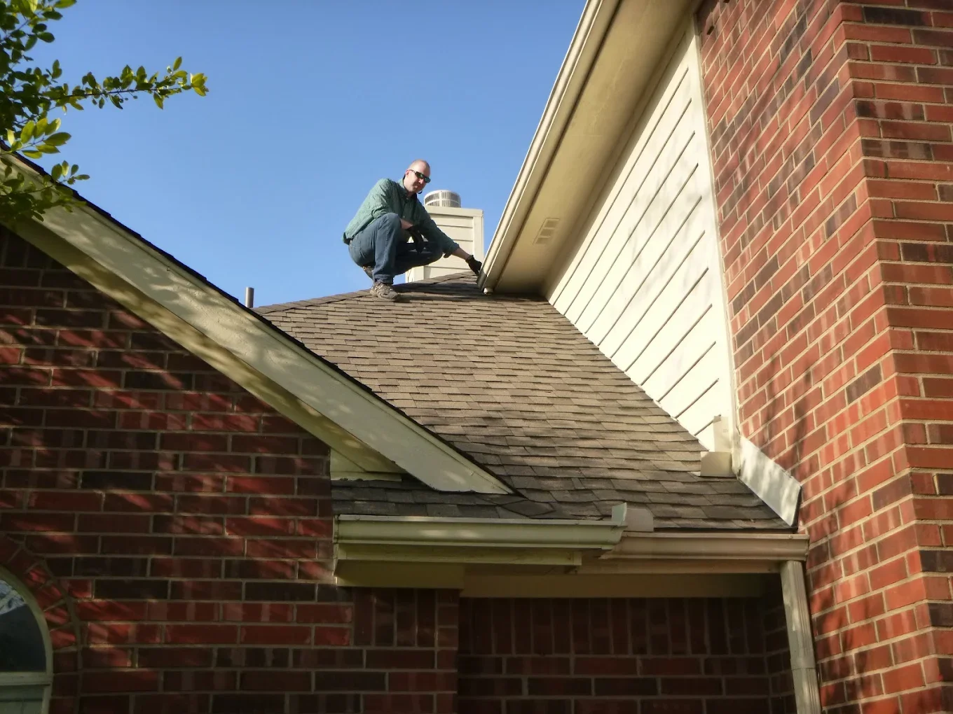 A man in sunglasses crouches on a roof, working on or inspecting the roof shingles near the chimney of a brick house under a clear blue sky.
