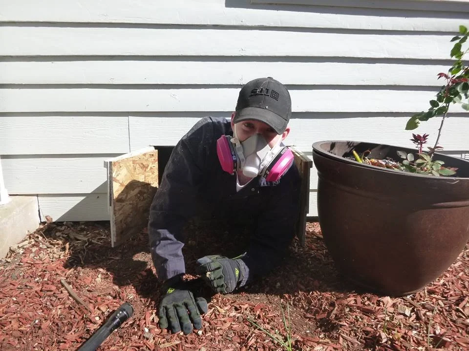 Person in work gloves and a face mask wearing a baseball cap, crouched next to a plant and a large flowerpot, working on a garden bed outside a house.