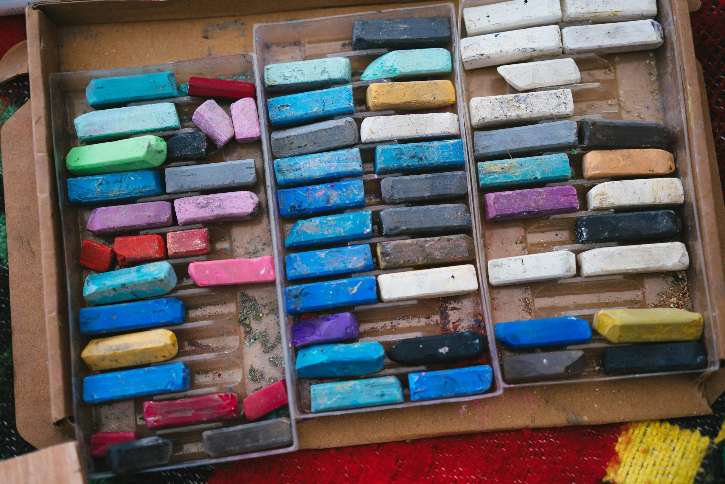A wooden box containing various colorful chalks, some with visible chalk dust and smudges, arranged in rows on a textured surface.