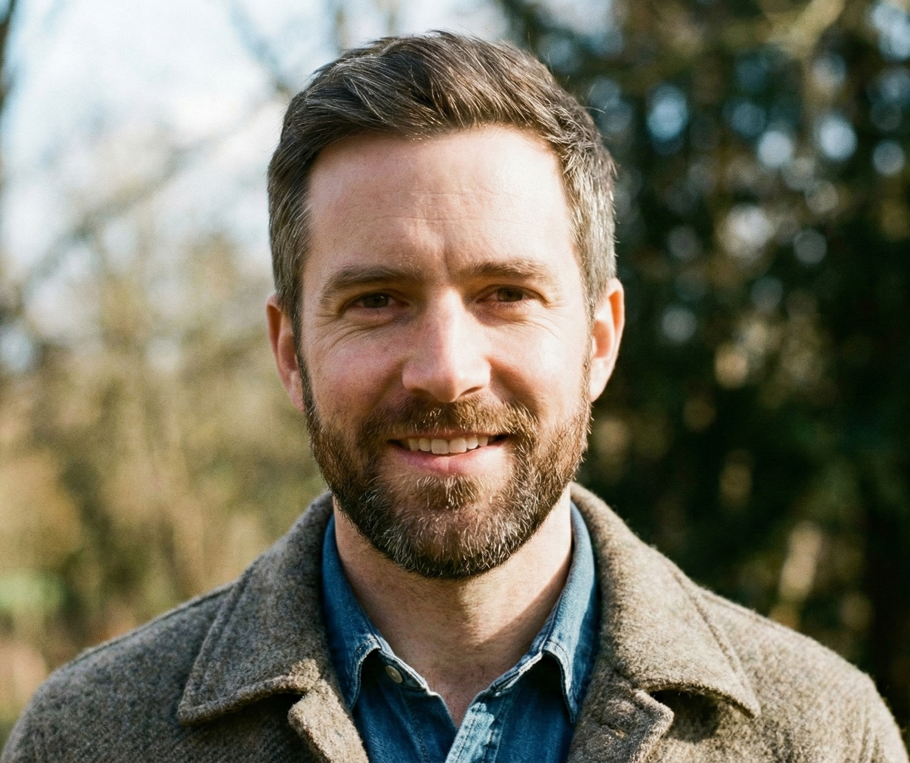 A smiling man with a beard and short dark hair, wearing a brown coat and a blue shirt, standing outdoors in a natural setting with trees in the background.