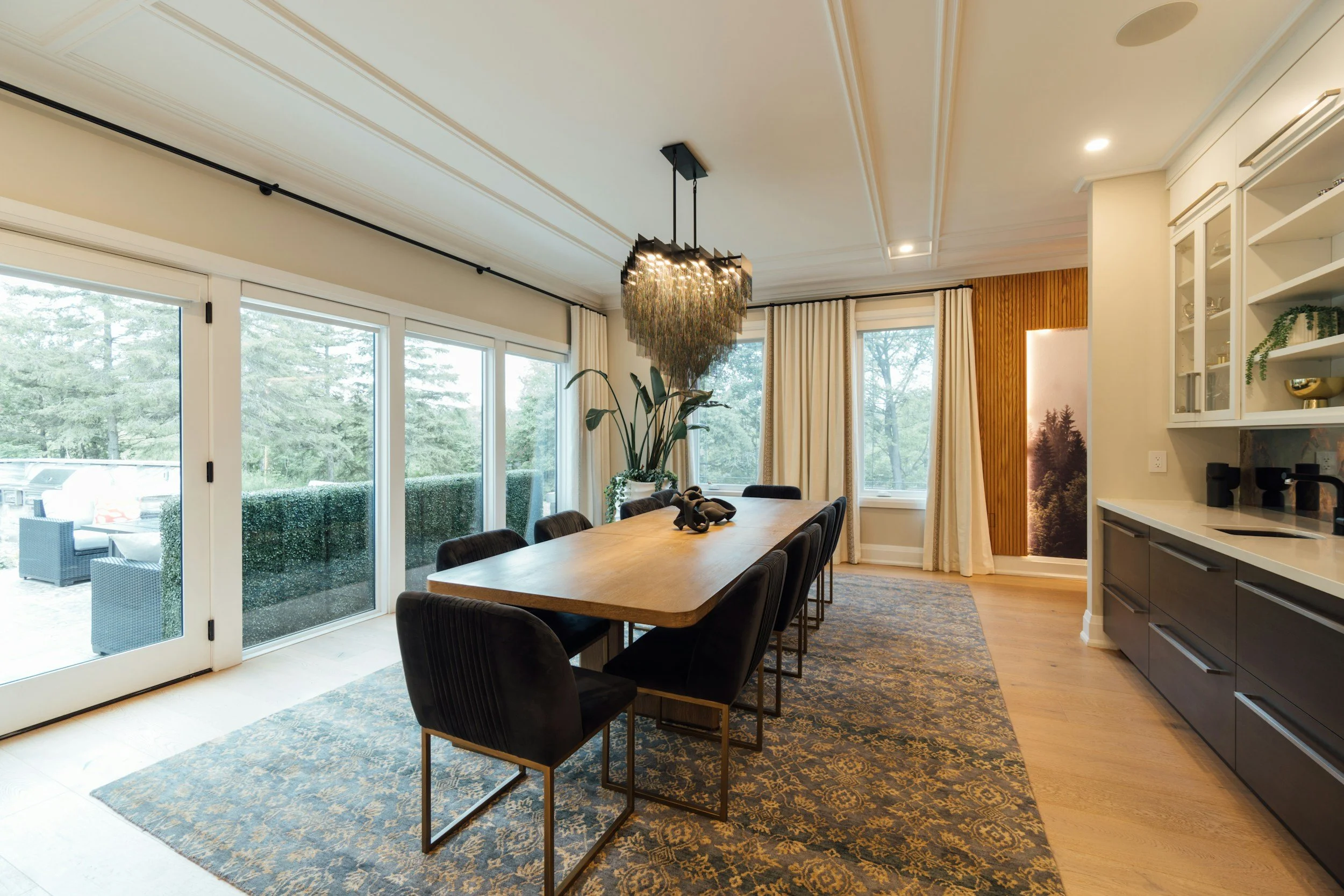 Dining room with a wooden table, black chairs, a modern chandelier, large windows, curtains, a large plant, and a sideboard with glass-front cabinets.