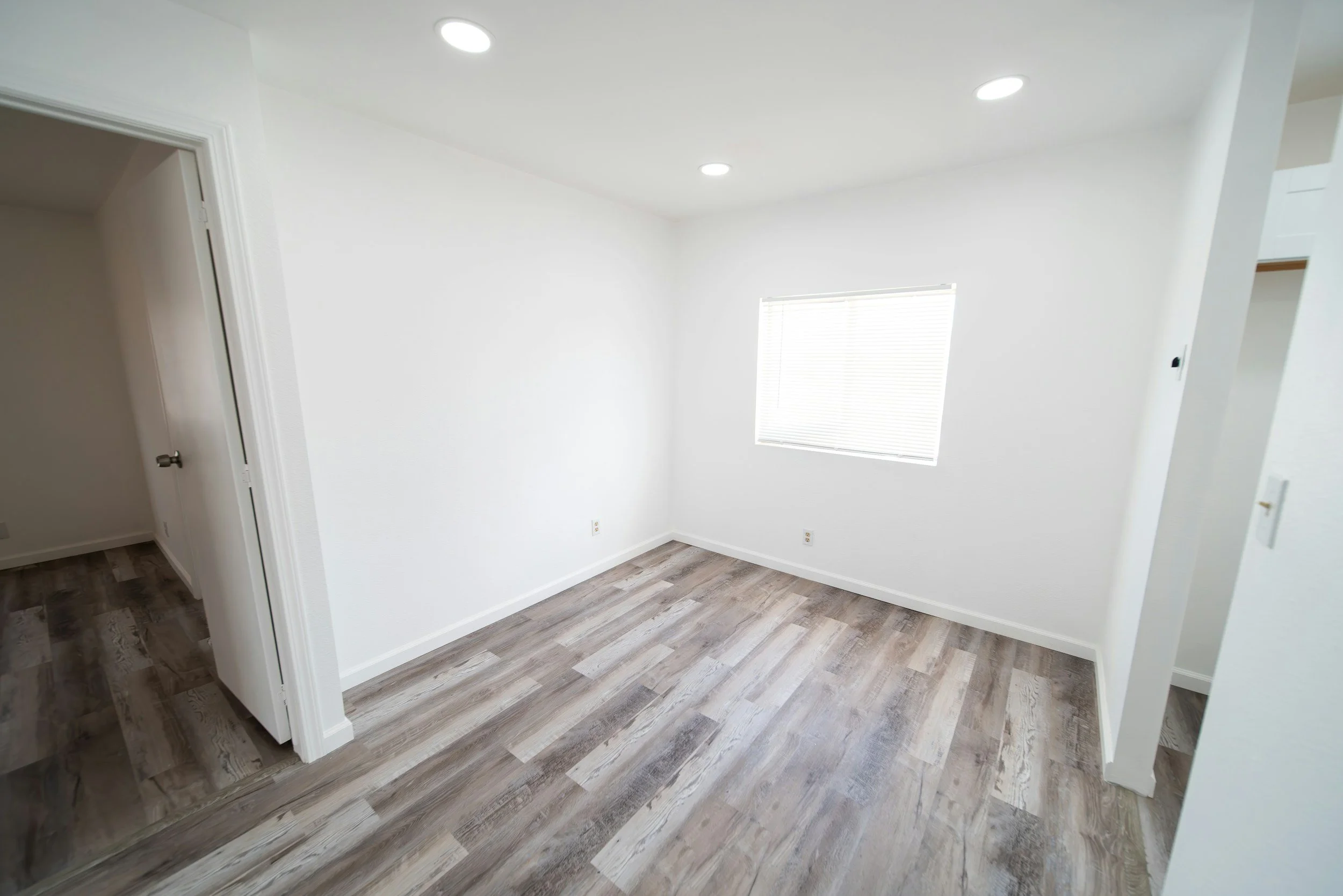 Empty white room with wood flooring, a window with blinds, and ceiling lights.
