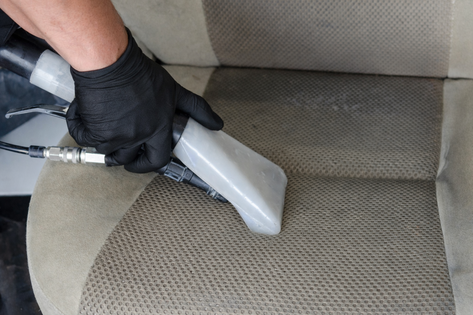 Person wearing black gloves using a handheld upholstery cleaning tool to clean a car seat with beige and brown fabric.