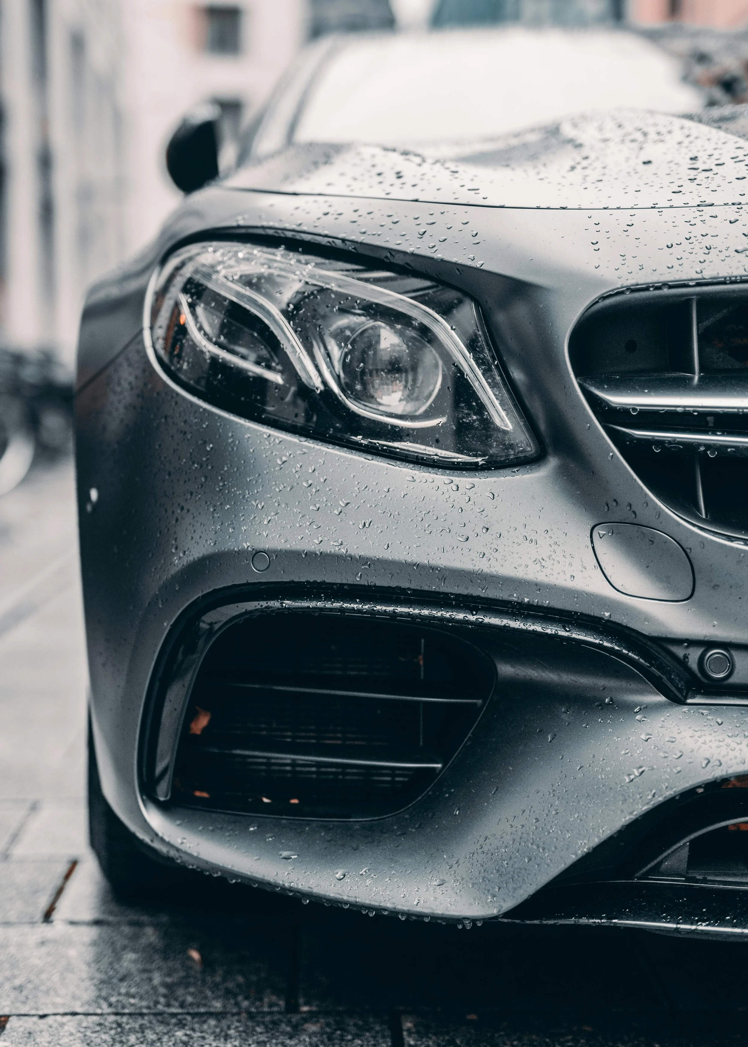 Close-up of the front left side of a gray luxury car with raindrops on the hood, showing the headlight and front grille.