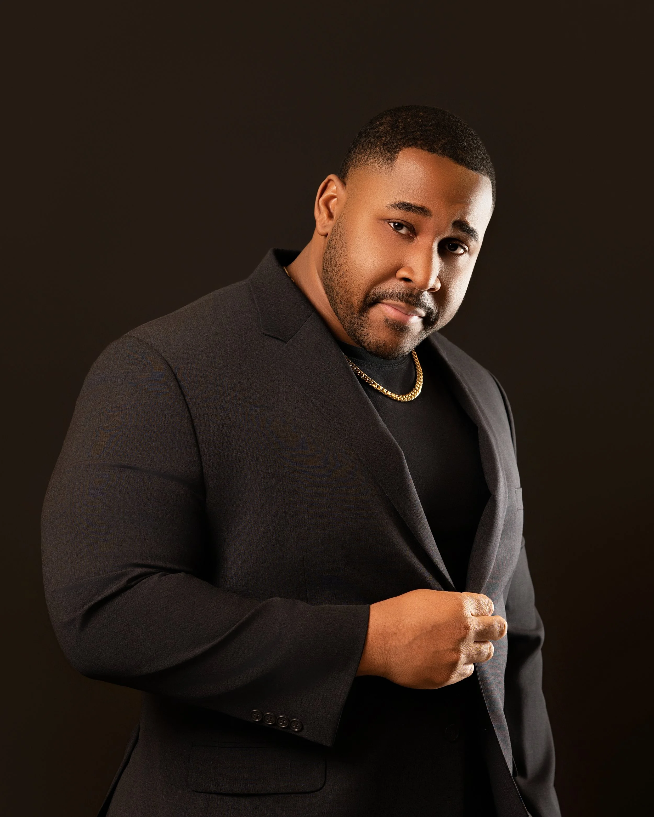 A man dressed in a black suit and black shirt, wearing a gold chain, posing confidently against a dark background.