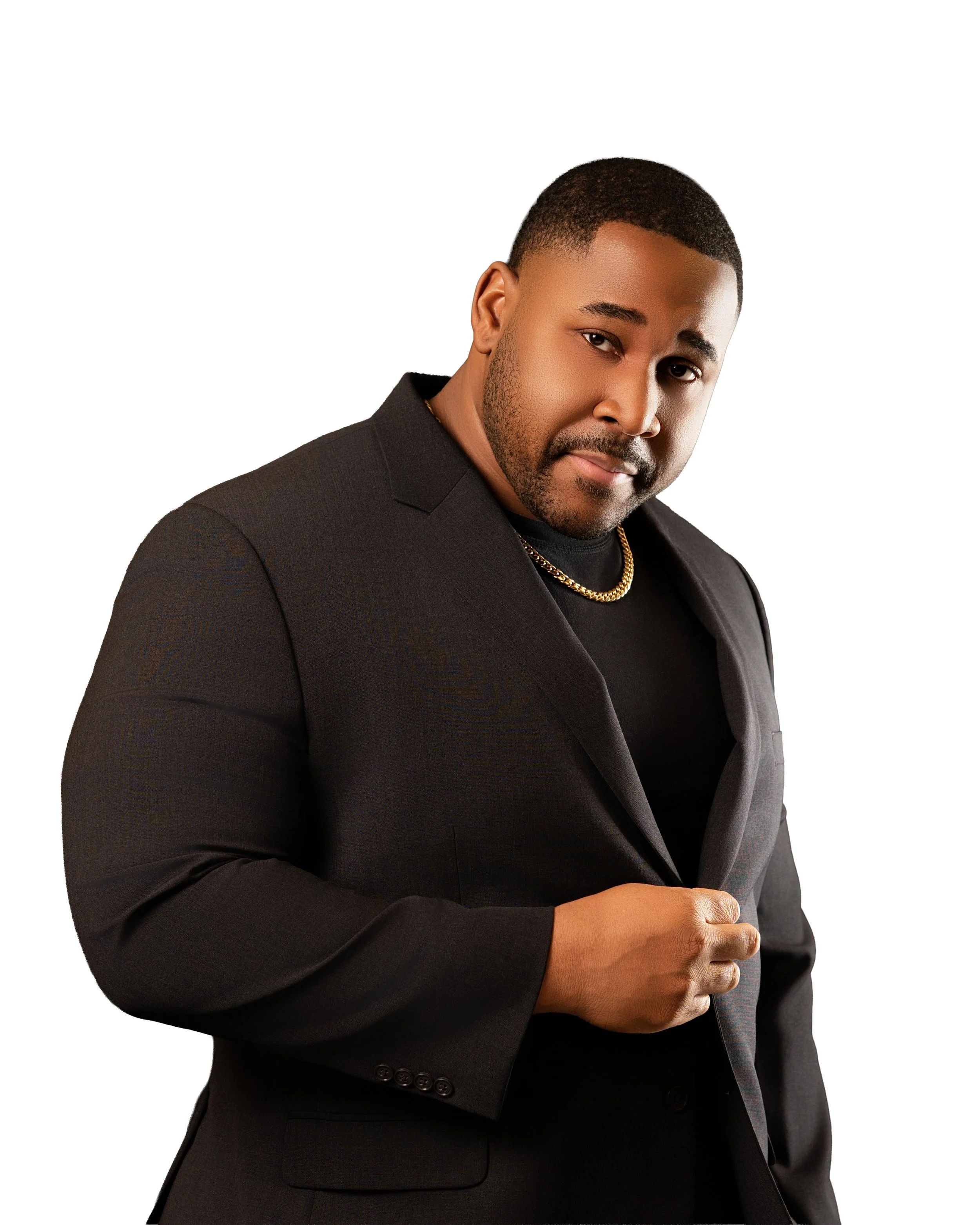 A man with a short haircut and beard dressed in a black blazer and black shirt, wearing a gold chain, looking directly at the camera with a confident expression.
