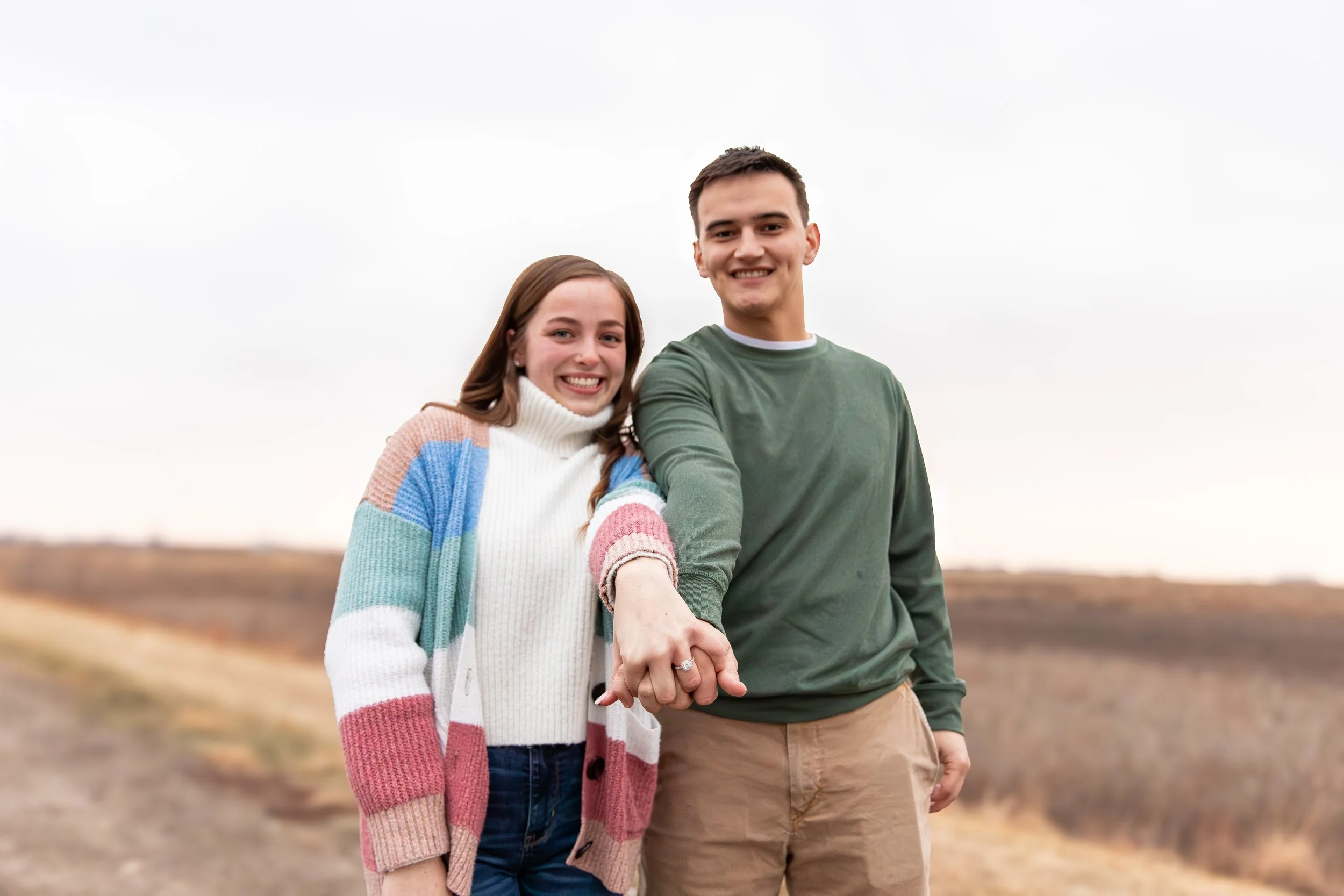 A smiling young woman and a smiling young man holding hands outdoors on a cloudy day.