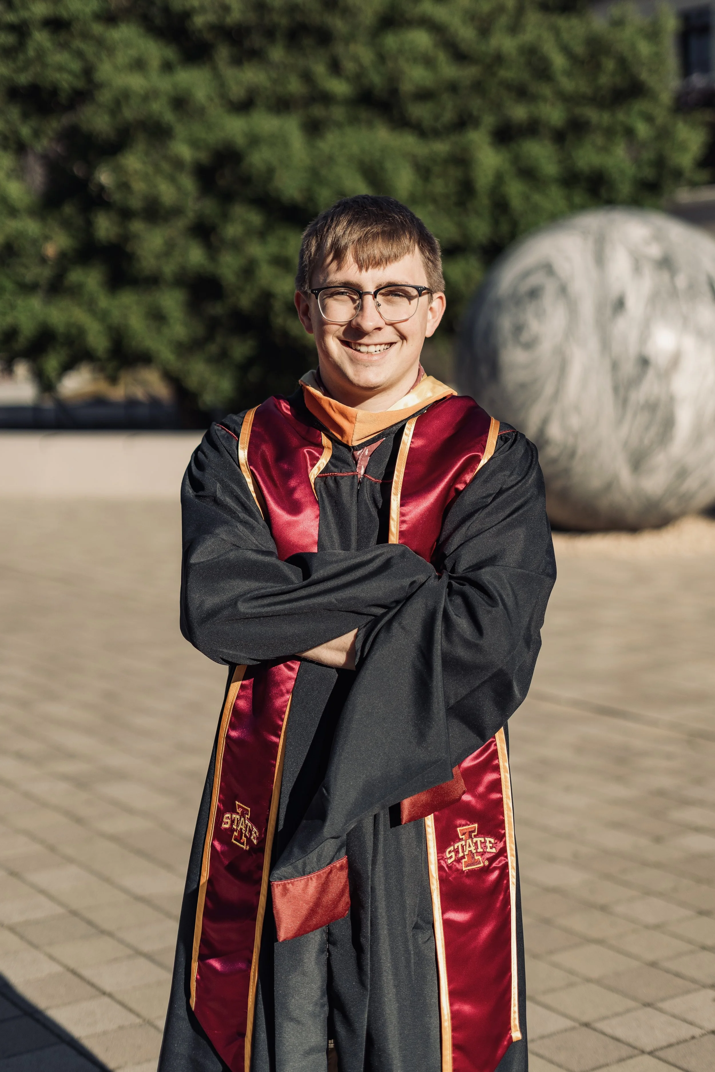 Stanford Campus Graduation Photo - A young man in a graduation gown and sash standing outdoors with arms crossed, smiling. Background includes a large bush and a spherical sculpture.