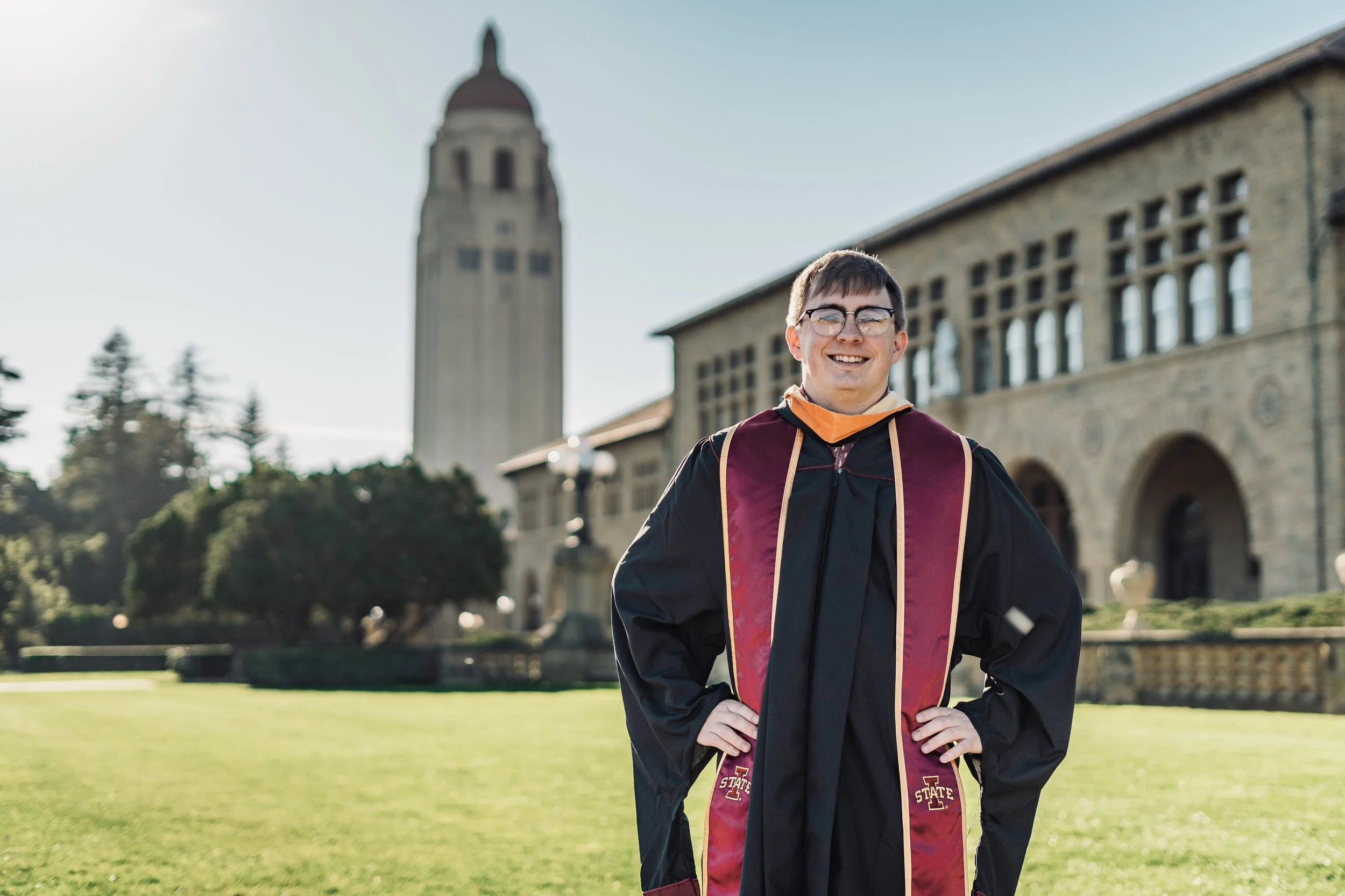 Stanford Campus Graduation Photo - Young man smiling in graduation gown and cap standing outdoors with university building and clock tower in the background.