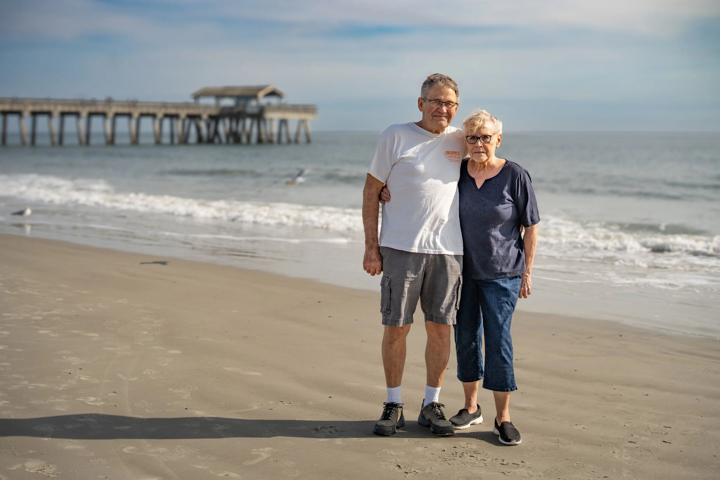 Family Photography at Beach - An elderly man and woman standing together on a sandy beach with ocean waves behind them and a pier in the distance. The man wears a white t-shirt and shorts, and the woman wears a dark t-shirt and shorts. 