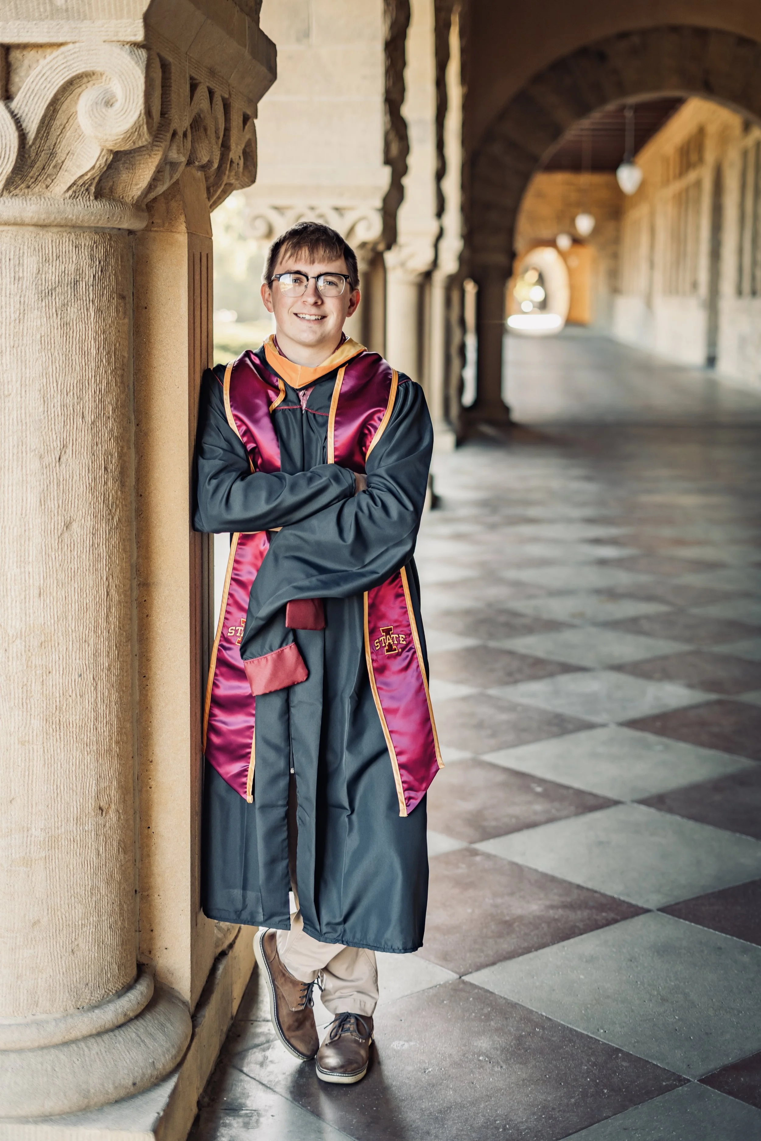 Stanford Campus Graduation Photo - A young man wearing glasses, a graduation gown, and a sash, leaning against a stone column with crossed arms, standing in a covered walkway with arches and checkered floor.
