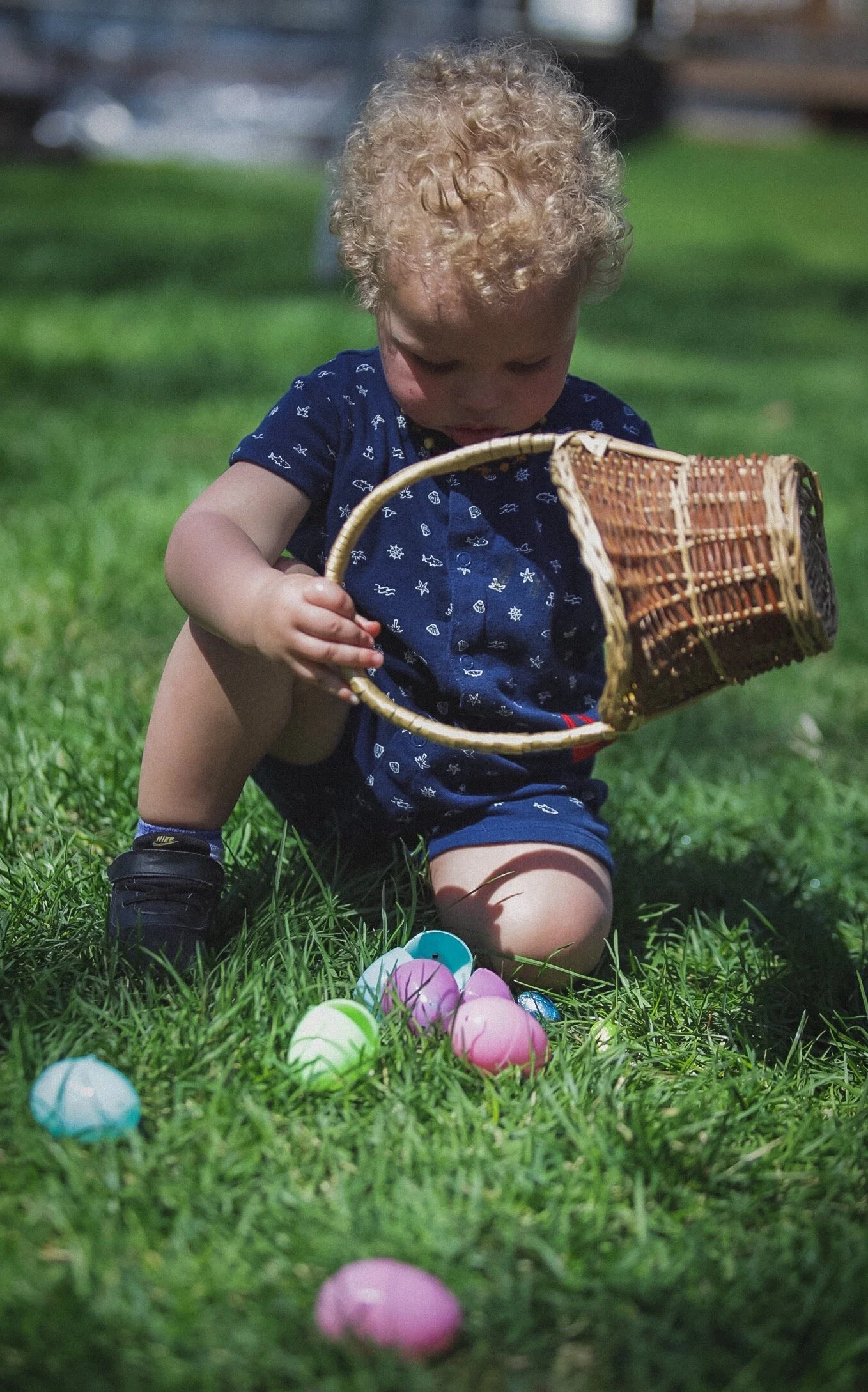 Family Photography with Kids - A young child with curly blonde hair wearing a navy blue shirt with white pattern and black shoes kneeling on the grass, holding a small basket, surrounded by colorful plastic Easter eggs.