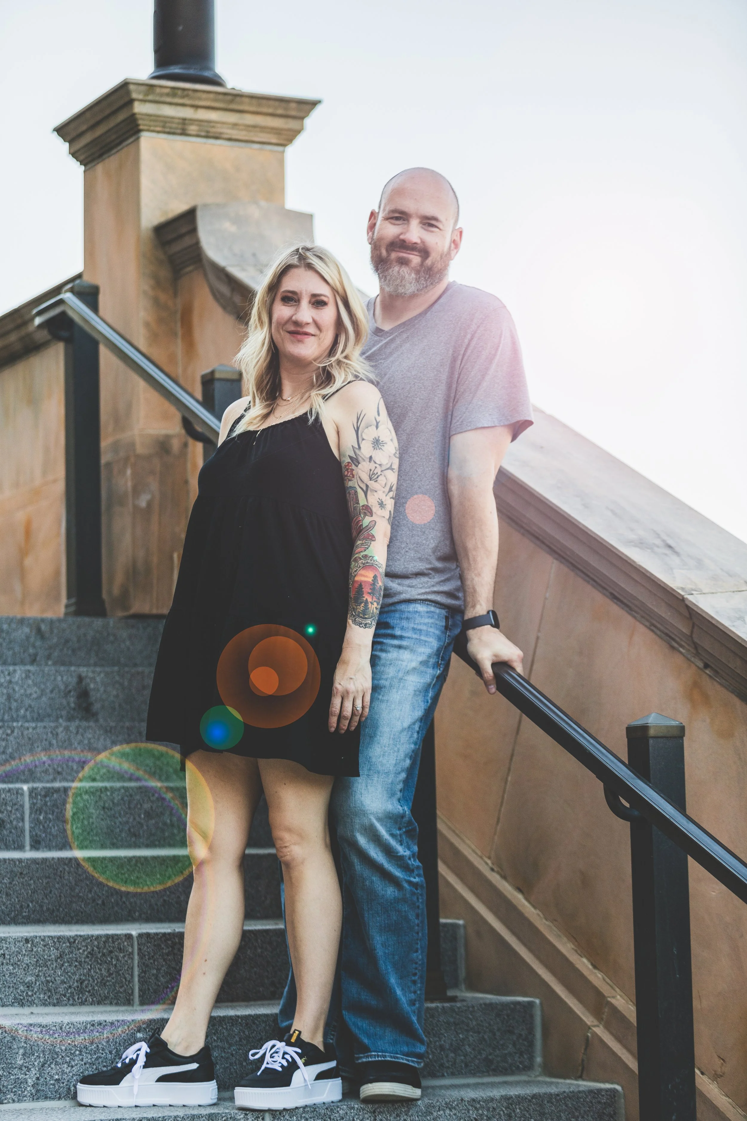 A smiling couple standing on outdoor stairs. The woman has blonde hair, tattoos on her arm, and is wearing a black dress and sneakers. The man has a beard and is wearing a gray t-shirt and jeans. The background includes a building with a decorative s