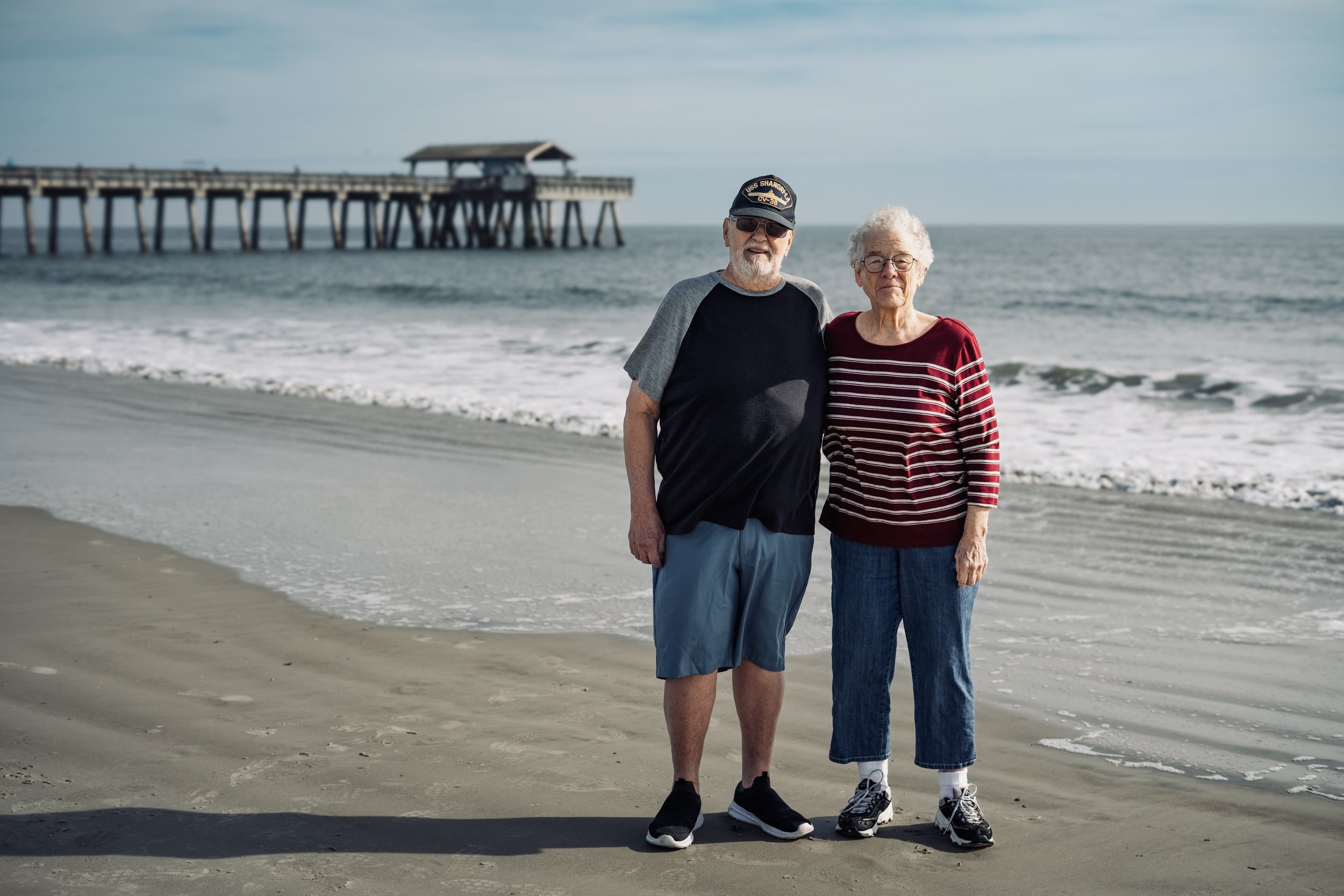 Family Photography at Beach - An elderly couple standing on the beach near the ocean, smiling and posing for a photo, with a pier in the background.