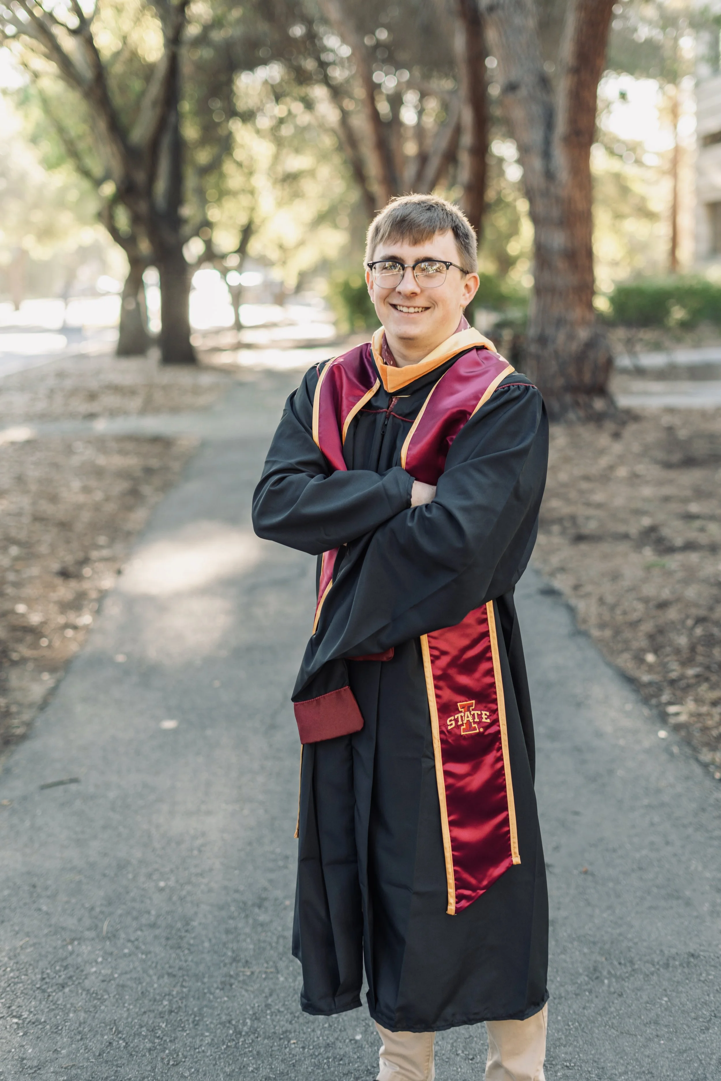 Stanford Graduation Photos - Young man in college graduation gown and cap standing on a sidewalk in a park, smiling, with trees and sunlight in the background.