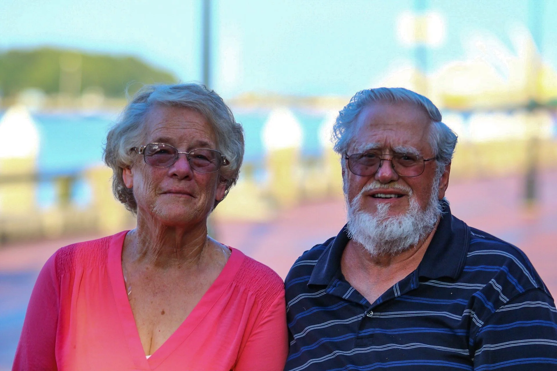 Family Photography/Couples Photos - An elderly woman with gray hair and glasses wearing a pink top, standing next to an elderly man with a beard, glasses, and gray hair wearing a dark striped shirt, outdoors at a sports field with blurred background.