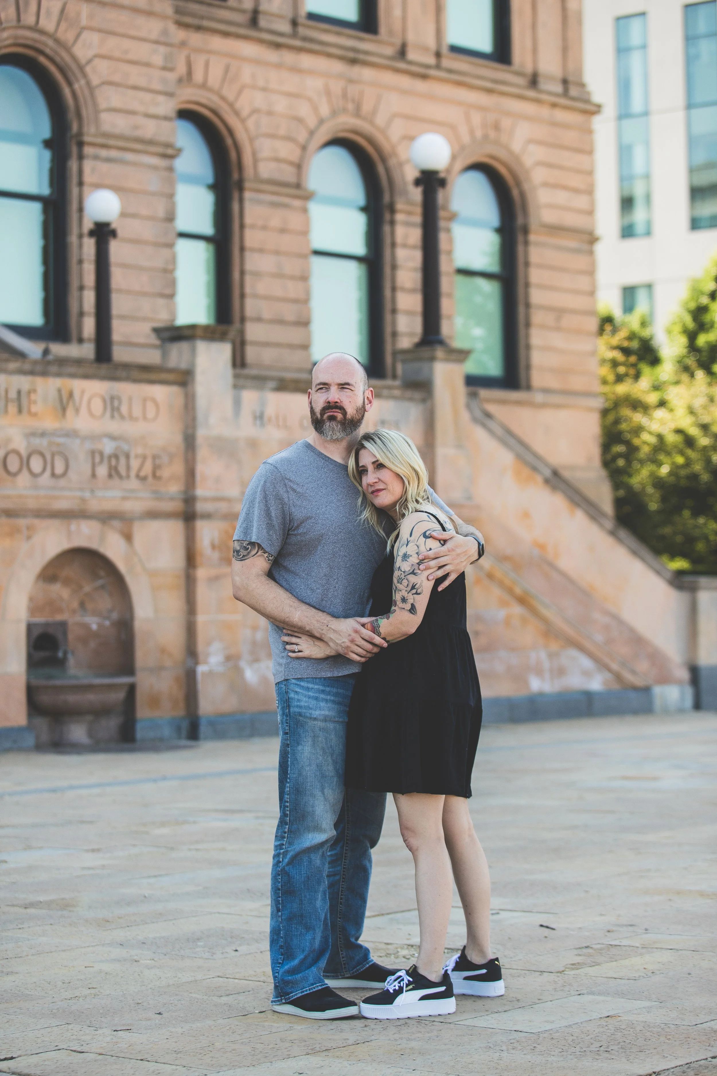 A couple stands close together outside a historic brick building with large arched windows, embracing each other with the man's arm around the woman's shoulders. The woman has blonde hair and tattoos on her arms, while the man is bald with a beard. T