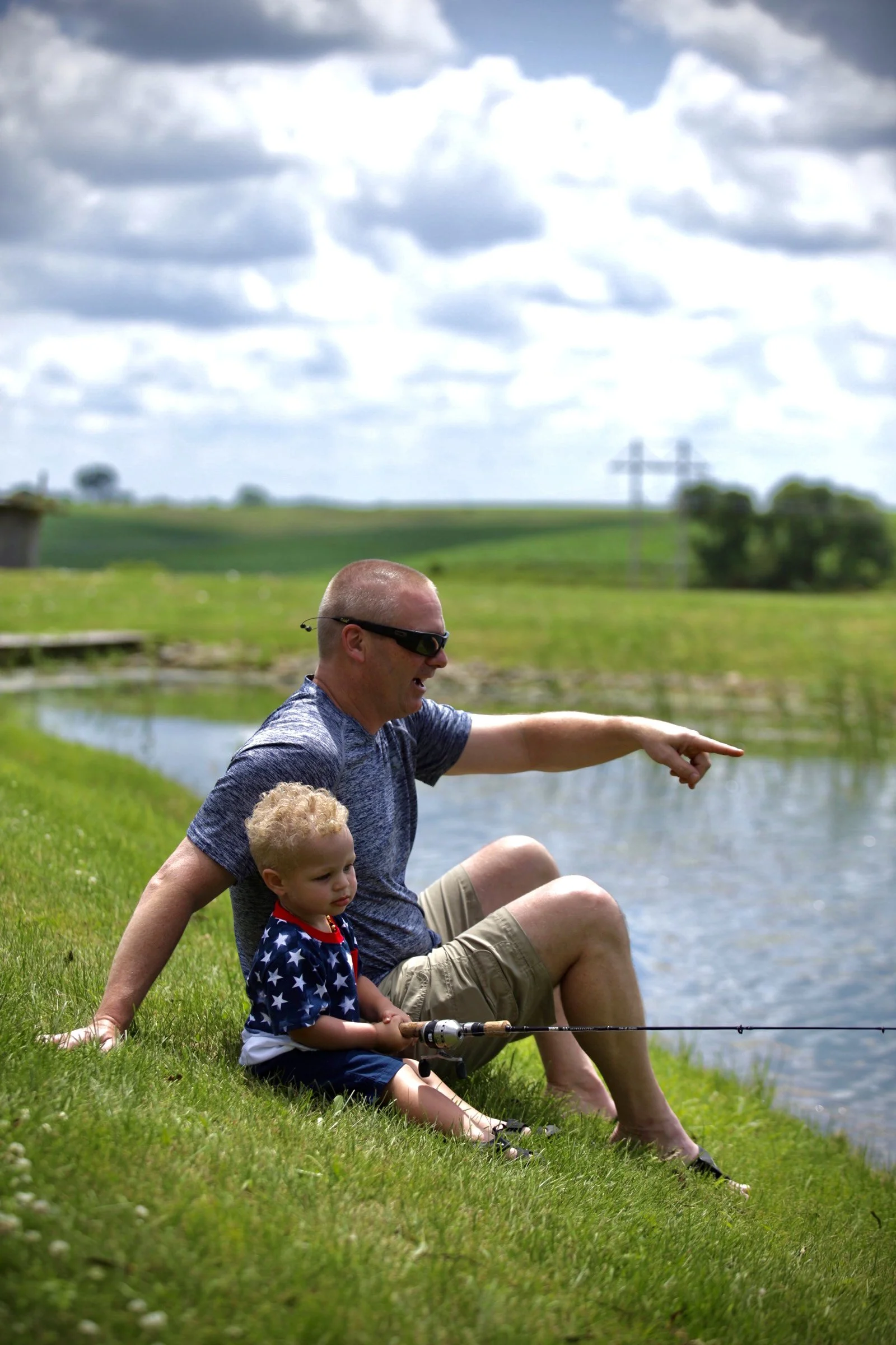 Family Photography with Kids - A man and two children sitting by a pond on a grassy bank, fishing with a dock and fields in the background under a cloudy sky.