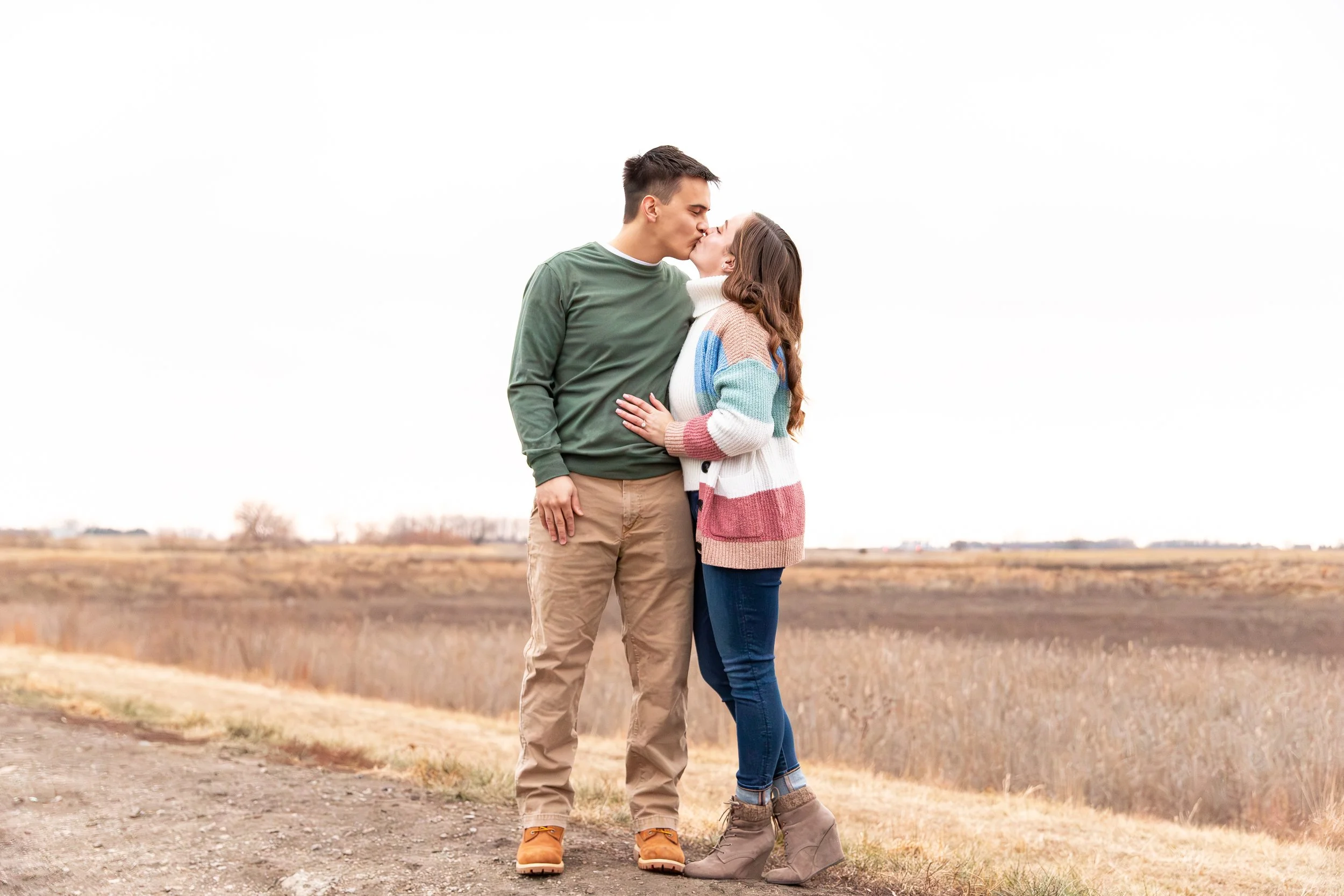 A young couple kissing outdoors in a large open field during fall, with the woman wearing a multicolored sweater and the man in a green shirt and beige pants.