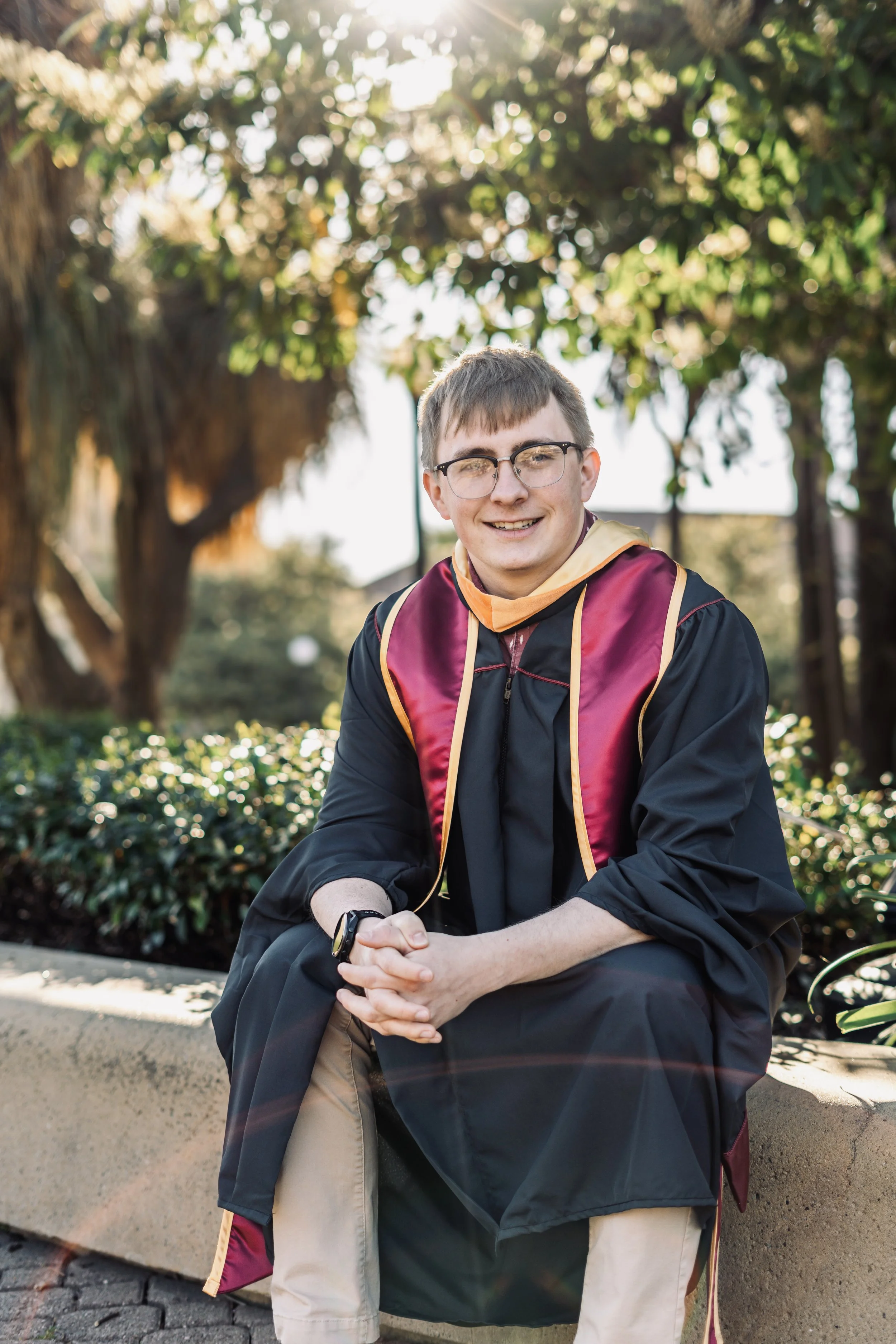 Stanford Graduation Photography - A young man in graduation cap and gown smiling outdoors, sitting on a paved sidewalk with greenery and large tree in the background.
