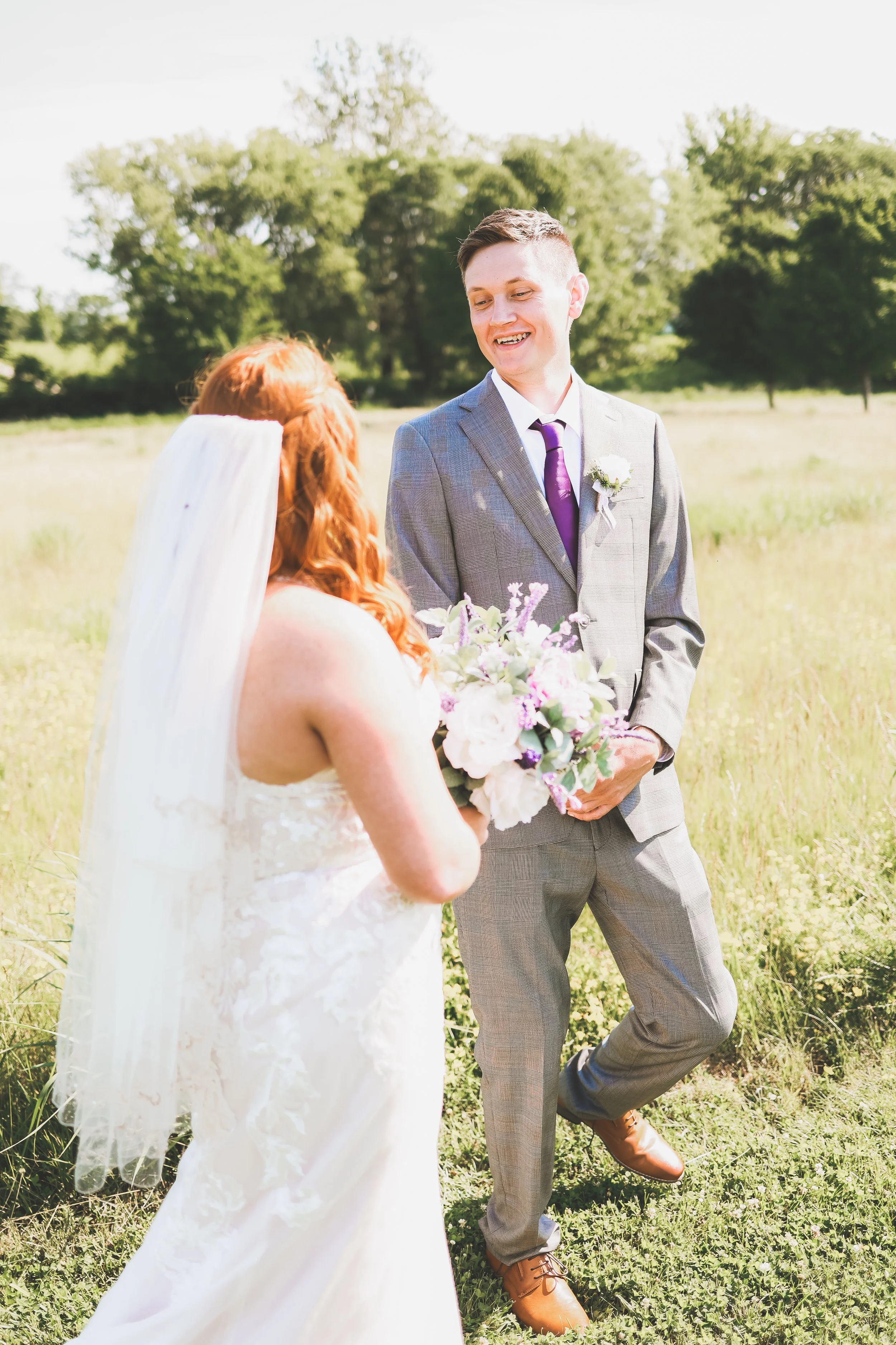 A wedding scene in a grassy field with trees in the background. A bride with red hair and a white veil and gown faces a groom with dark hair, wearing a gray checked suit with a purple tie and a white boutonniere. The bride holds a bouquet of flowers.