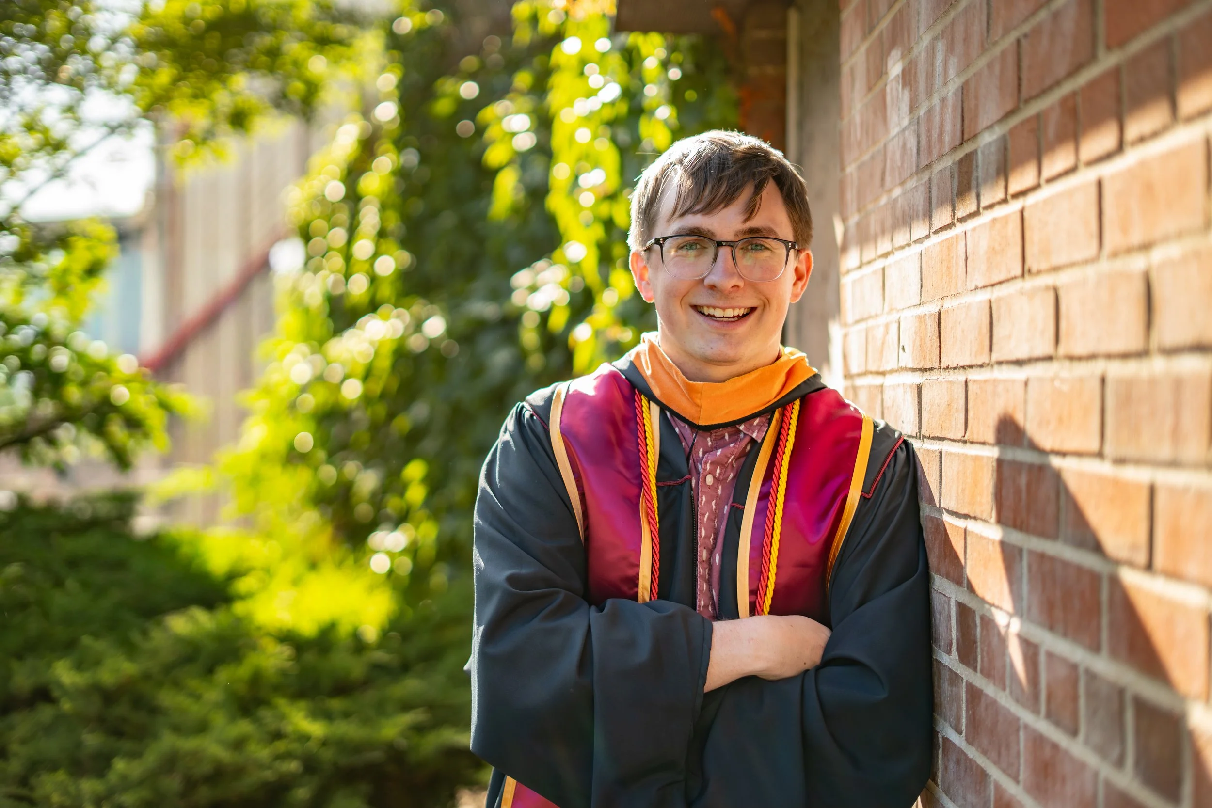 Senior Graduation Photo - A young man with glasses, wearing a graduation gown and cords, smiling and leaning against a brick wall outdoors with sunlight and green foliage in the background.