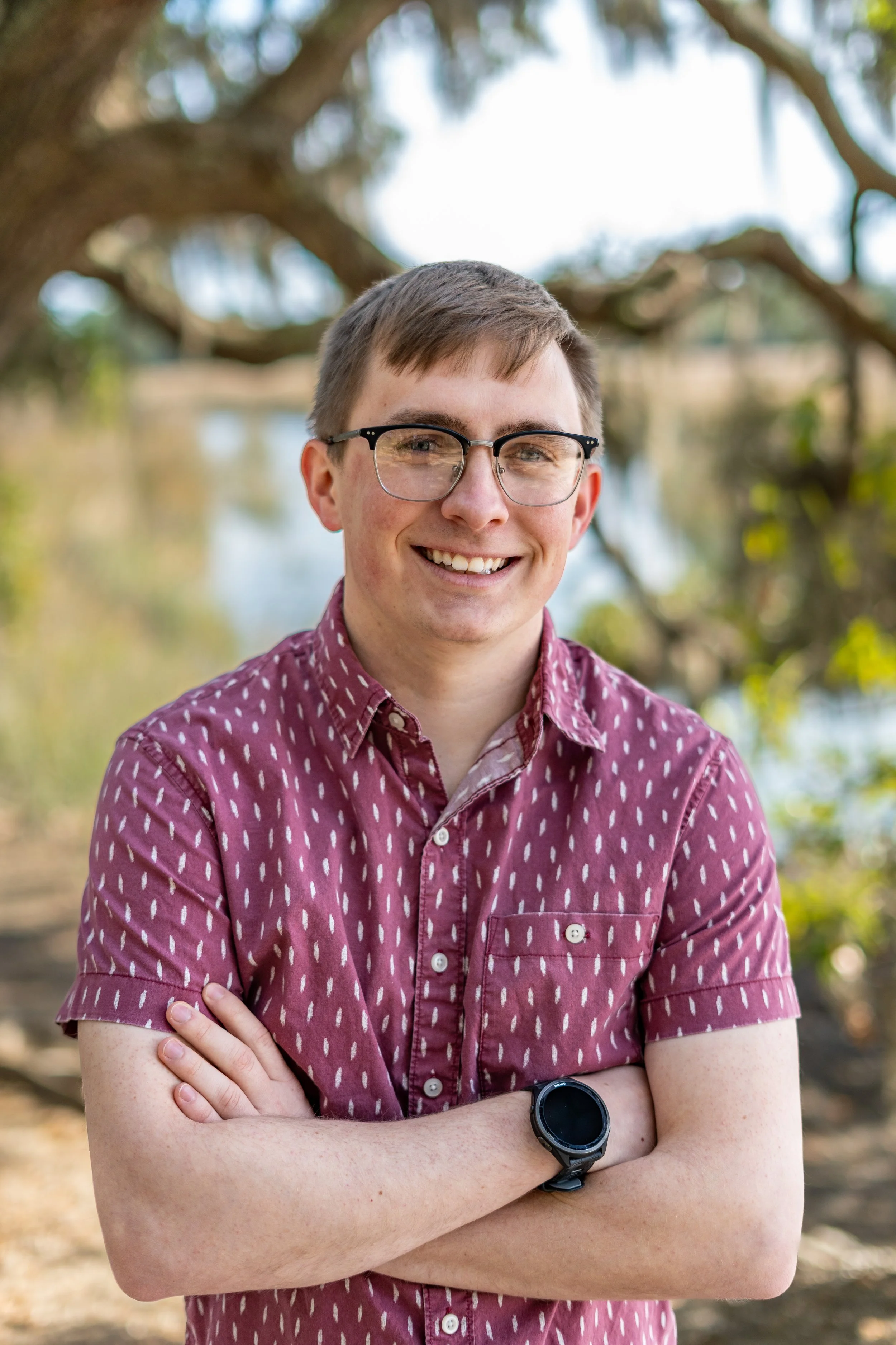 Professional Headshot - A young man wearing glasses and a maroon patterned shirt is smiling and standing outdoors with trees in the background.