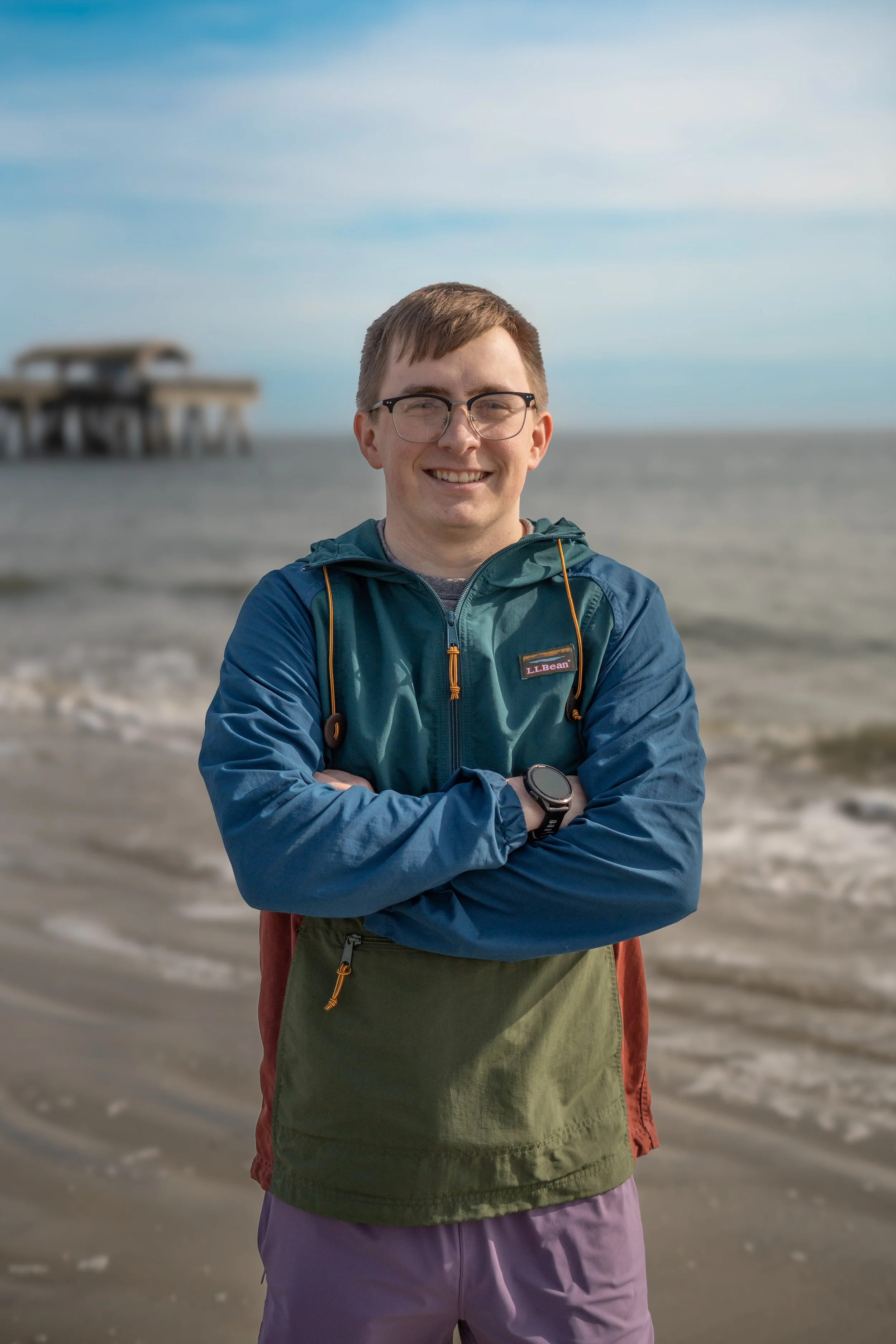 Family Portrait Photography - A young man with glasses smiling on a beach, standing with arms crossed, wearing a blue jacket and purple shorts, with a pier in the background under a partly cloudy sky.