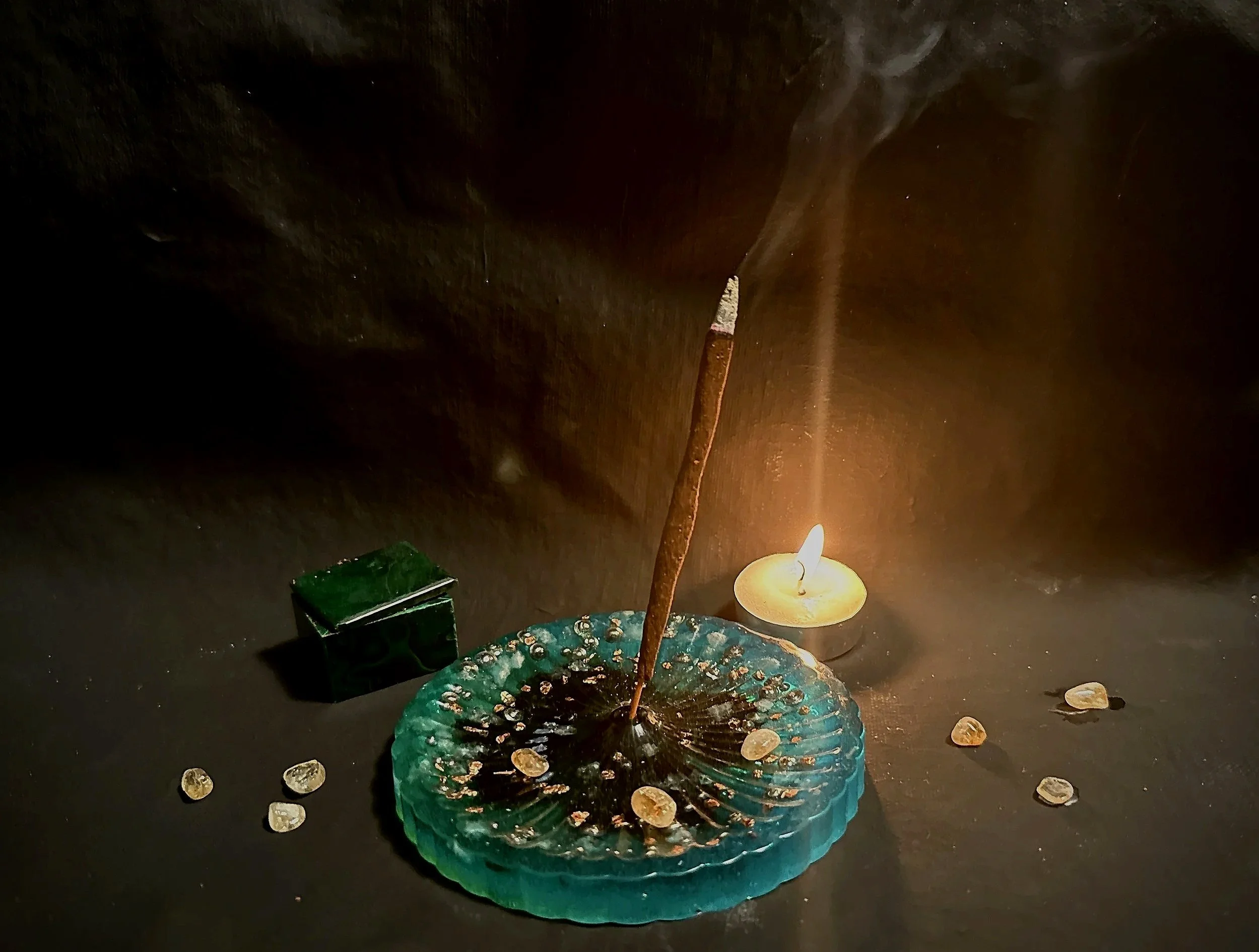 A lit candle  near an incense stick on a hand made resin incense burner, near a malachite box, surrounded by scattered crystals.