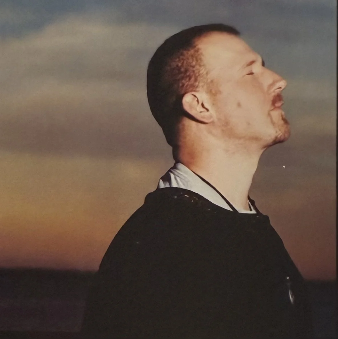 A man with closed eyes and a slight smile, facing right, stands outdoors during sunset or sunrise.