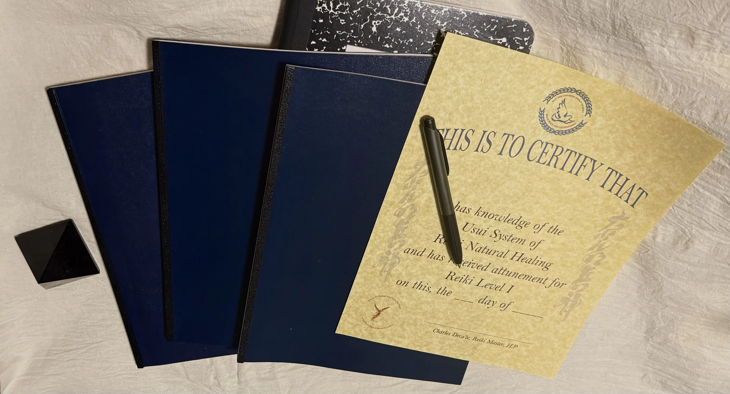 A set of three closed notebooks, a pen, and a certificate on a textured surface. The notebooks are dark blue and black with textured covers, while the certificate is yellow with printed text and a logo, partially visible.