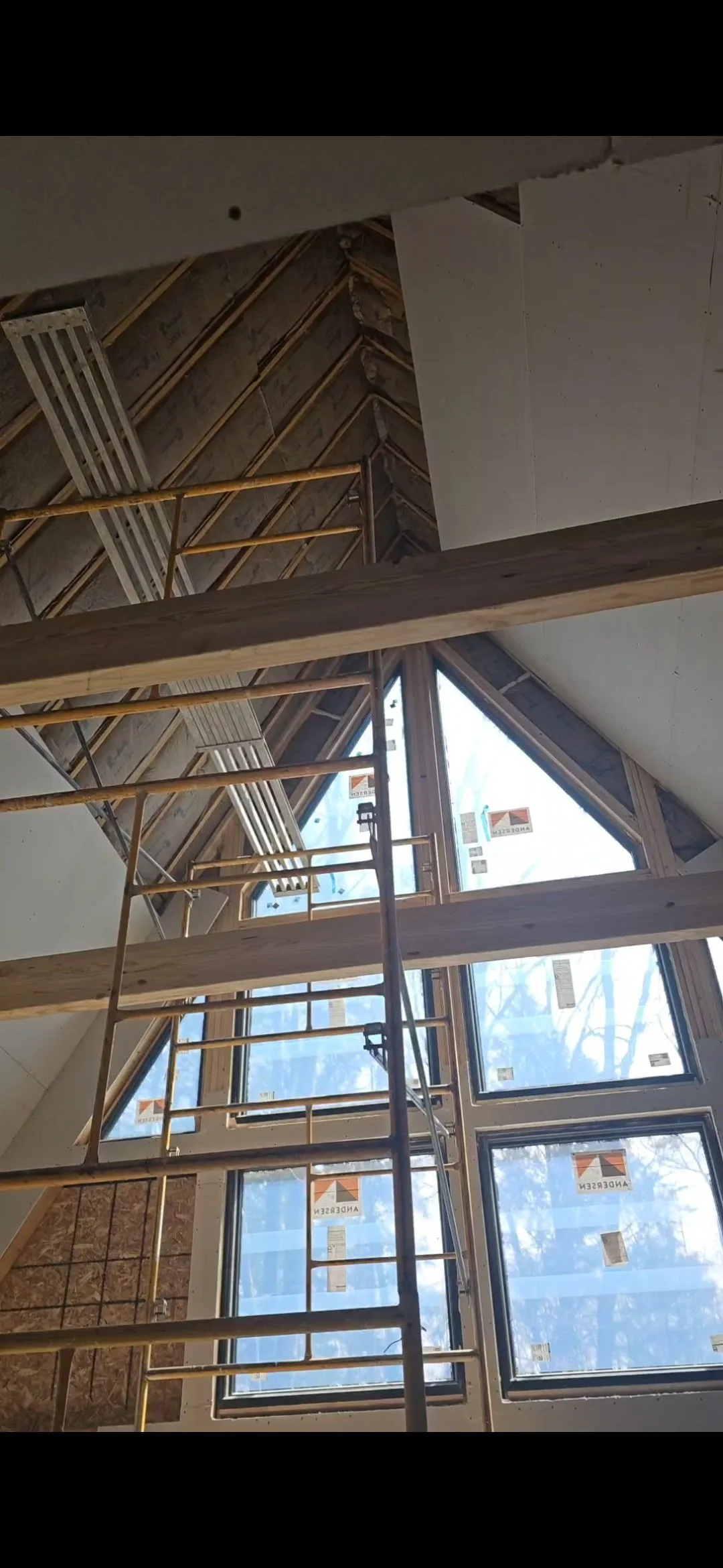 Interior en construcción con un escabel de madera y andamios de metal, ventanas grandes en forma de triángulo y rectángulo, estructura de madera en el techo parcialmente terminada.