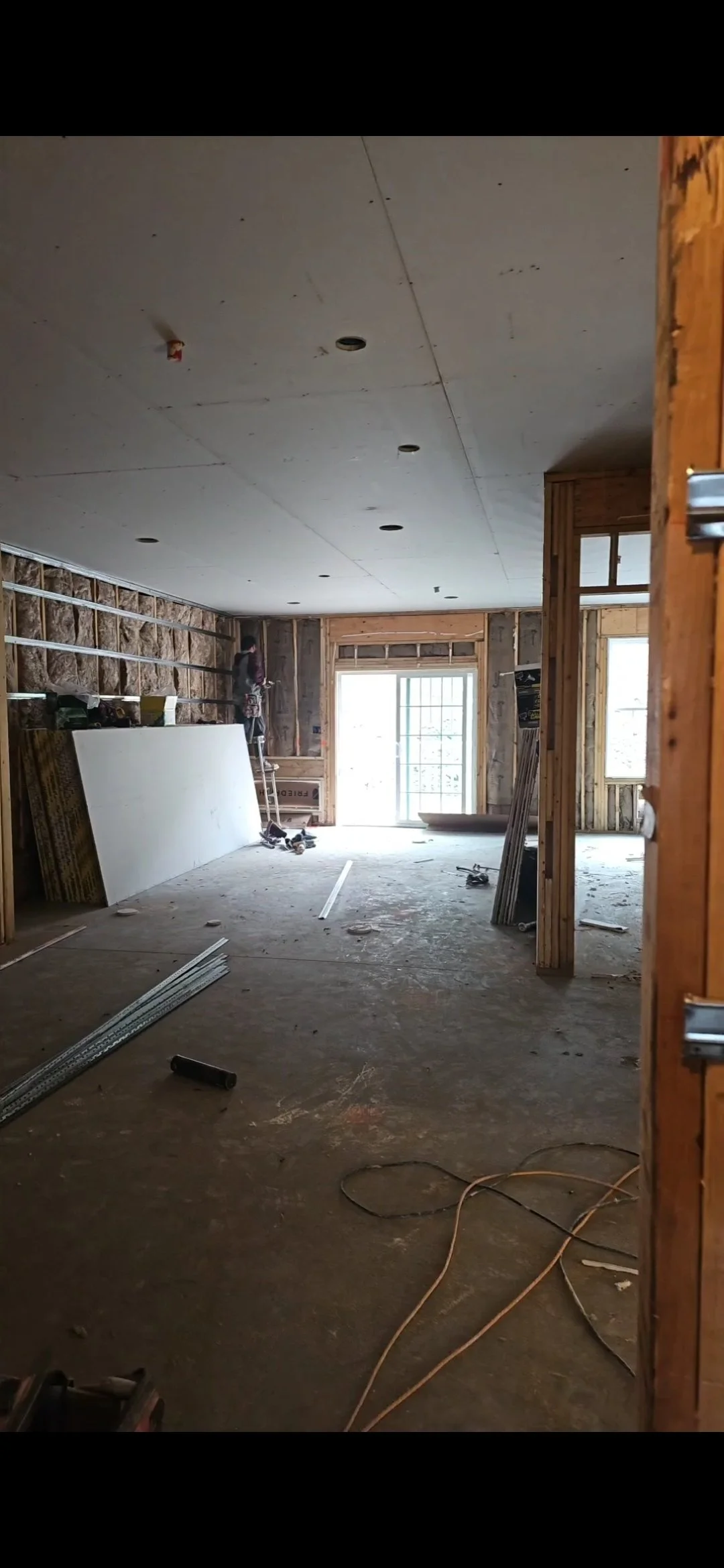 Interior de una casa en construcción, con paredes de madera, herramientas y materiales de construcción en el suelo, y un trabajador en escaleras cerca de una ventana grande.