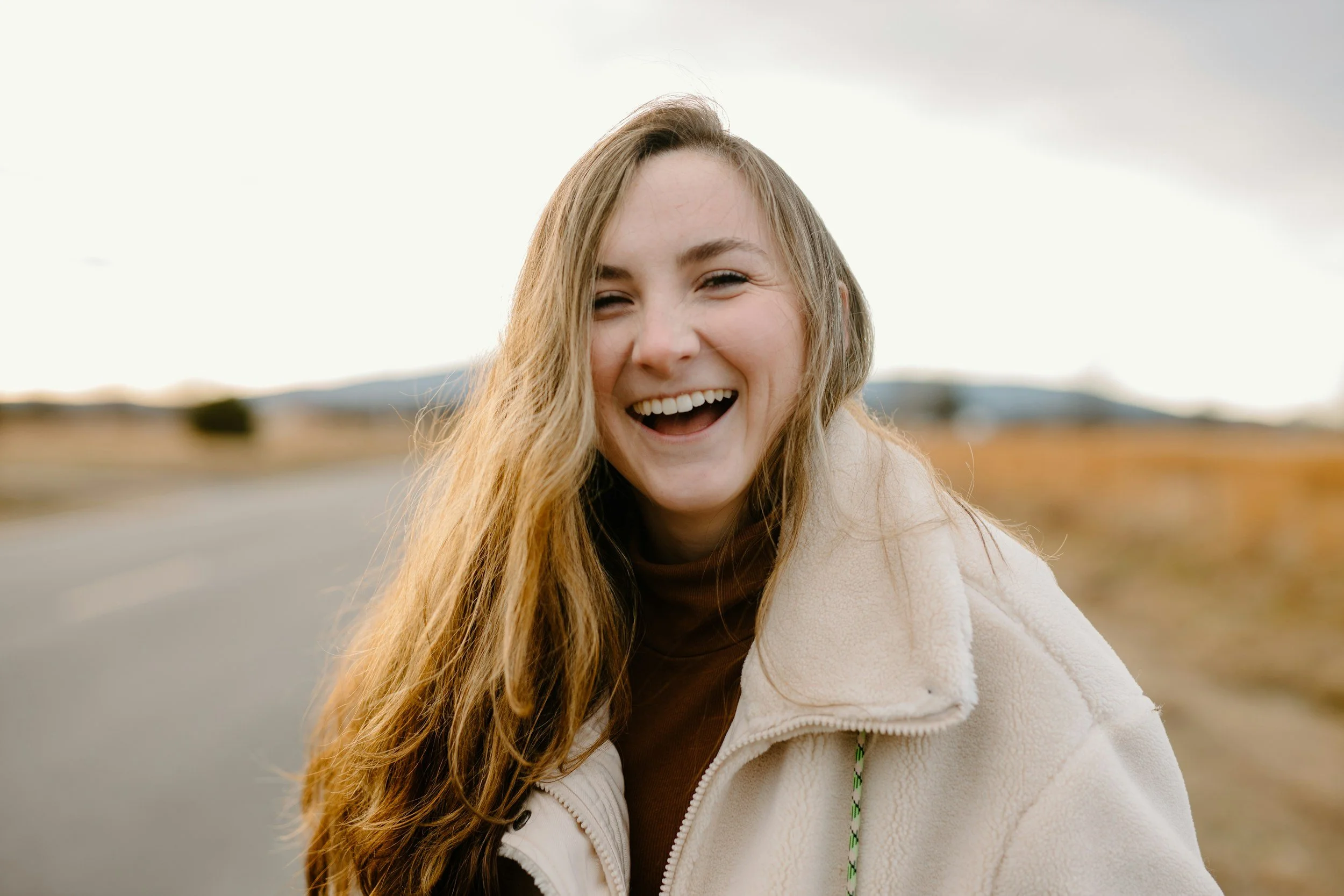 A young woman with long, wavy blonde hair smiling and laughing outdoors, wearing a cream-colored fleece jacket, with a blurred rural landscape in the background.