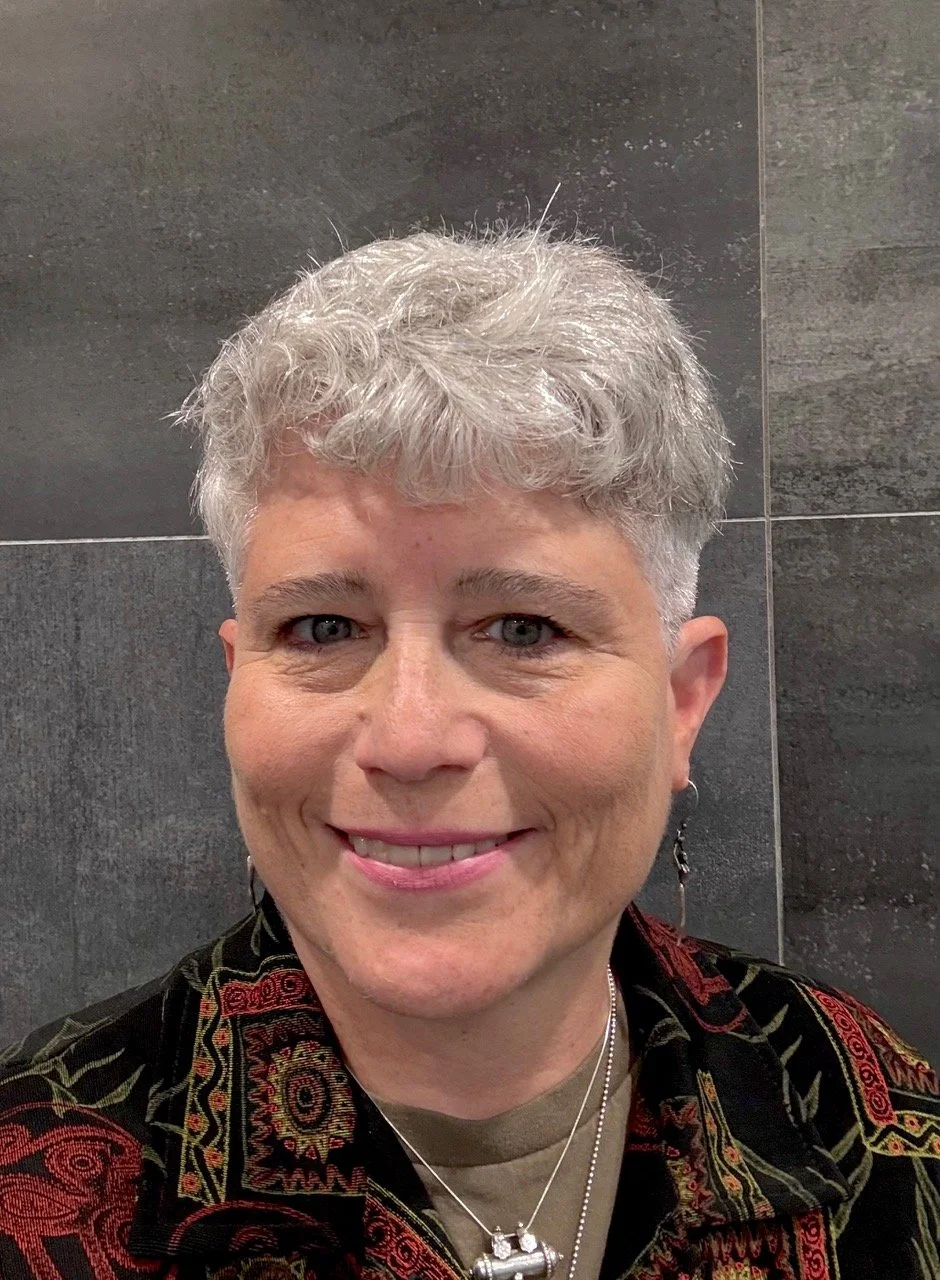 Close-up of a smiling person with short, curly gray hair, wearing earrings, a patterned jacket, and layered necklaces, standing against a dark tiled wall.