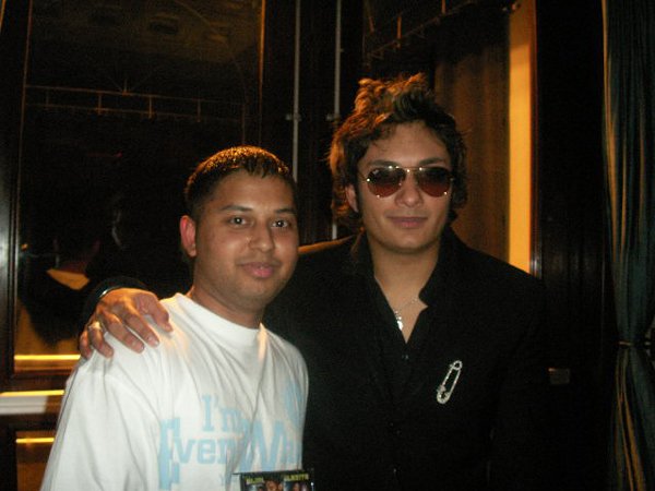 Two men standing together indoors, with the man on the right wearing sunglasses and a black shirt, and the man on the left wearing a white t-shirt, both smiling at the camera.