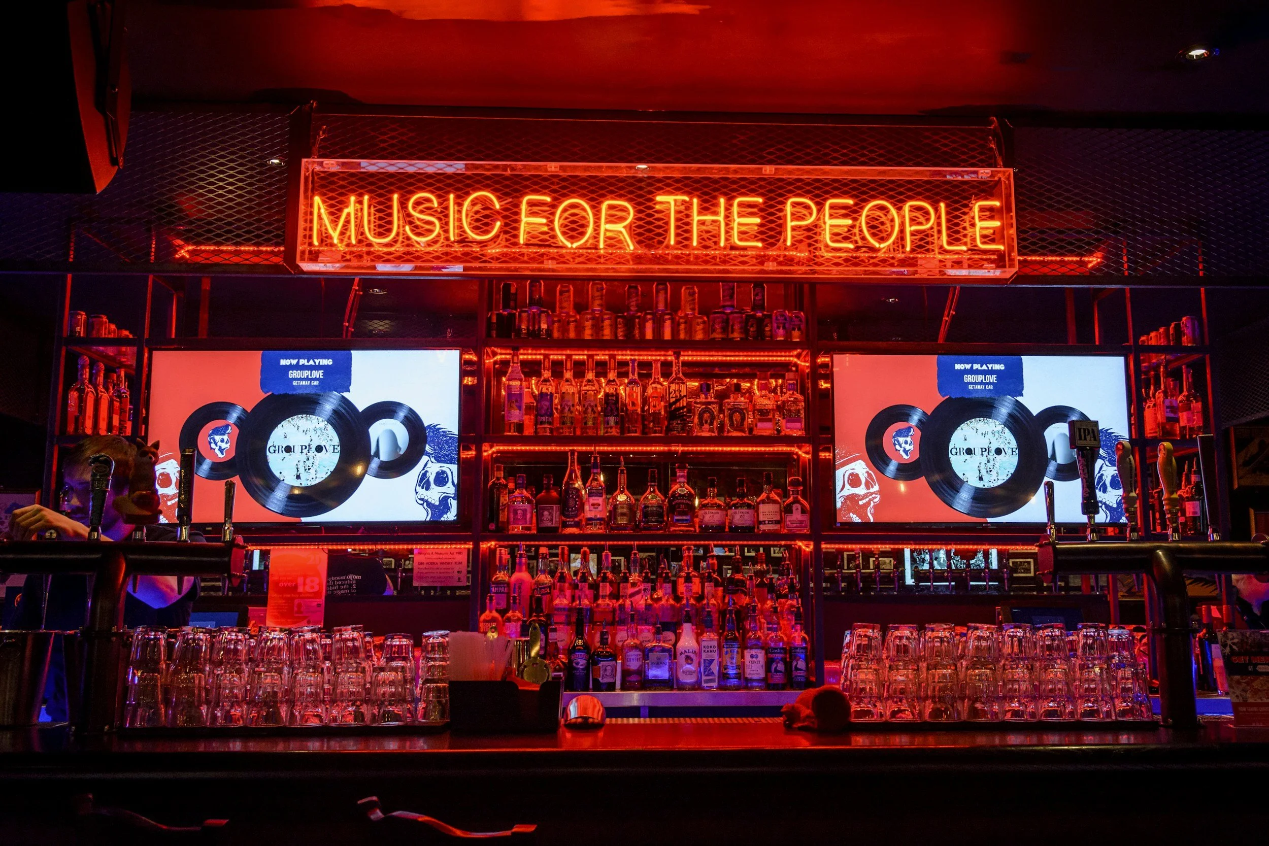 A bar with shelves of alcohol bottles, two TV screens displaying album covers, and a bright red neon sign reading 'MUSIC FOR THE PEOPLE'