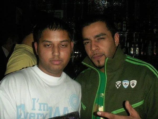 Two men posing for a photo indoors, one wearing a white shirt, the other in a green jacket with badges, in a dimly lit setting.