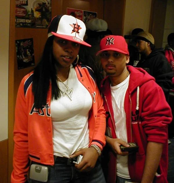 Two young adults posing together in a store, both wearing baseball caps and casual clothing. The woman is wearing a black and white Houston Astros cap, an orange and navy jacket, and a necklace. The man is wearing a red New York Yankees cap, a red hoodie, and appears to be holding a camera or small device.