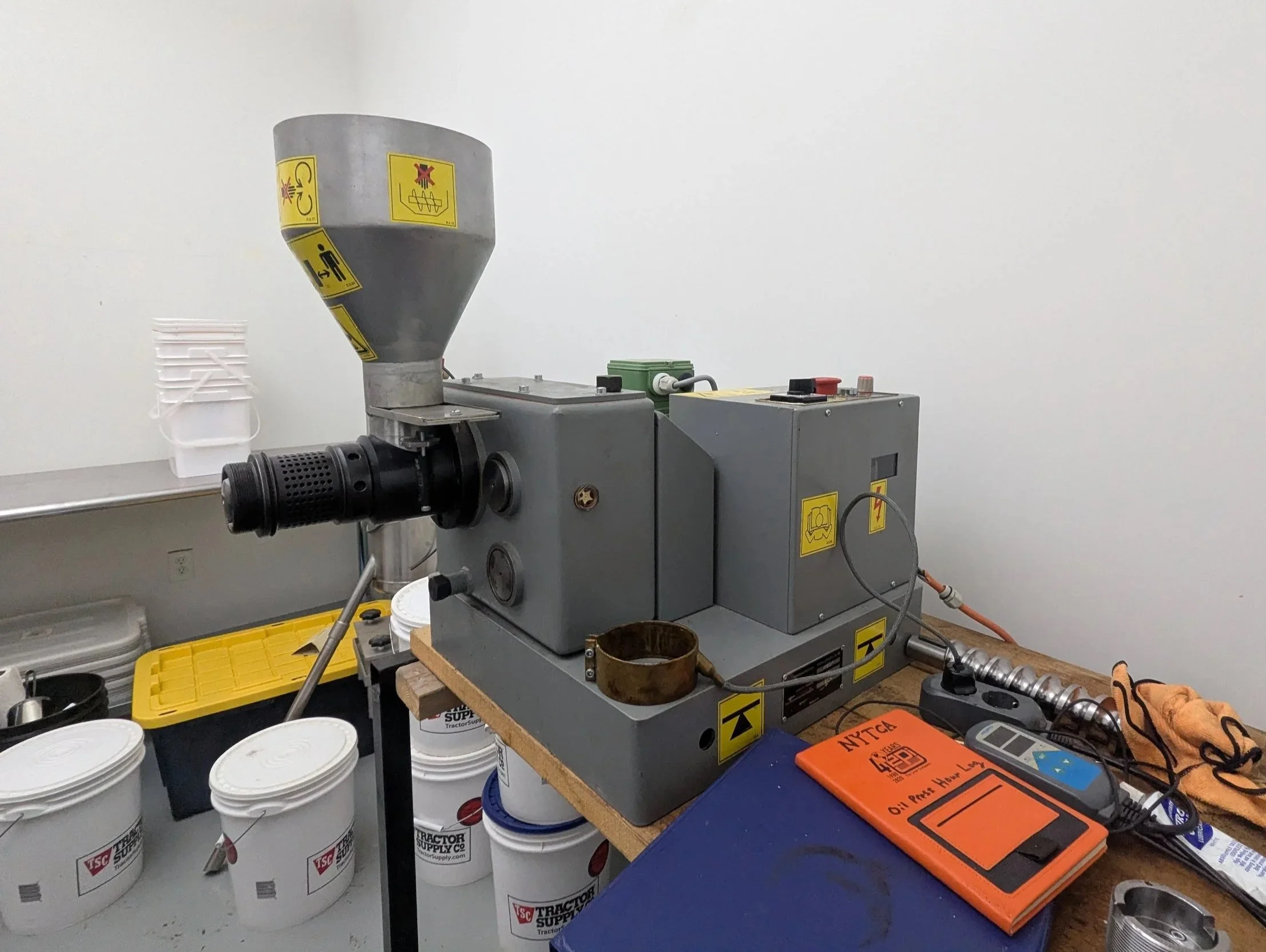 A metal machine with a funnel on top and a black handle on the front, situated on a workbench in a workshop. Several buckets and tools are nearby.