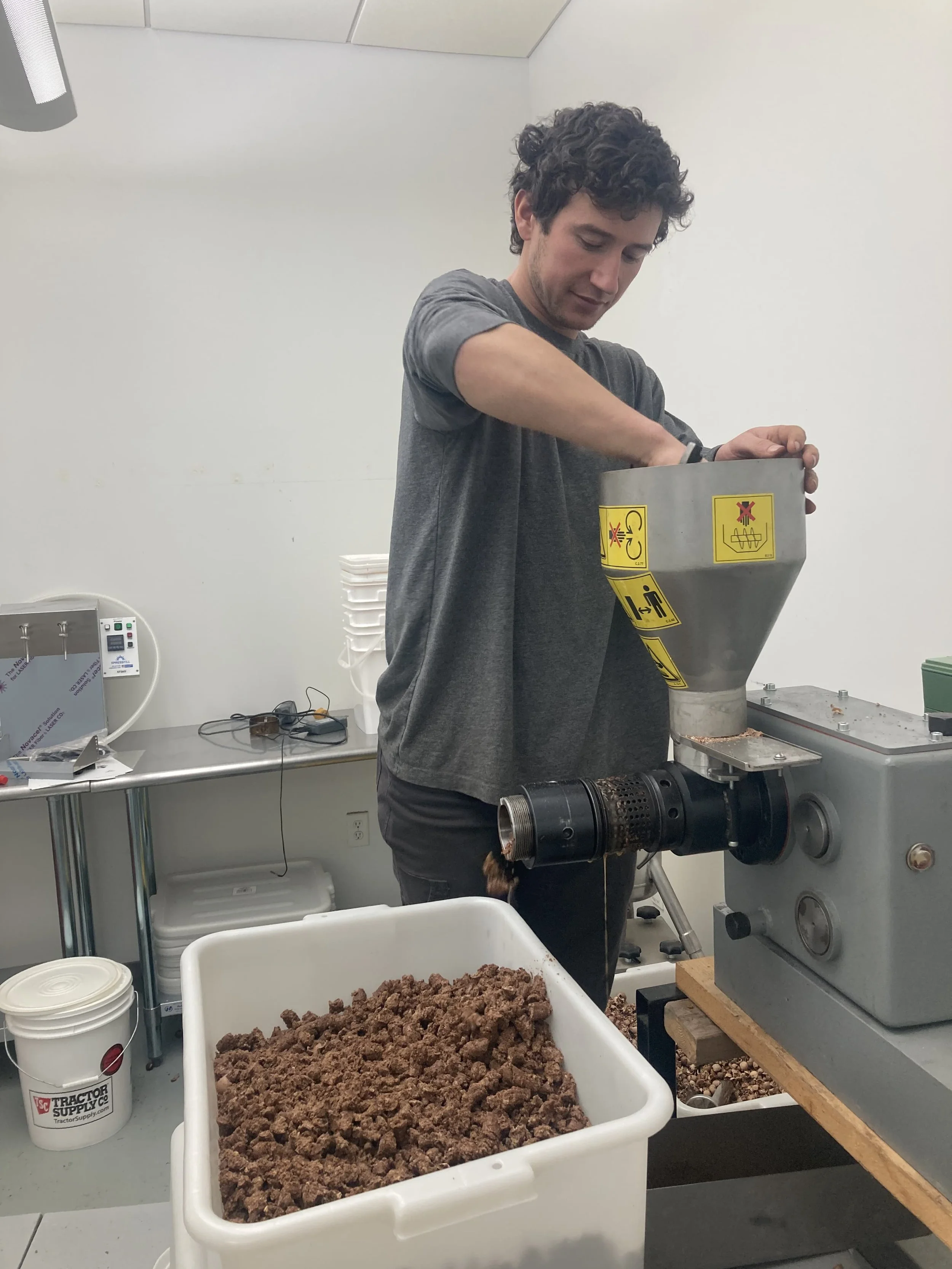 A man with curly dark hair operating a machine in a laboratory or industrial setting, with a large container of brown material in front of him.