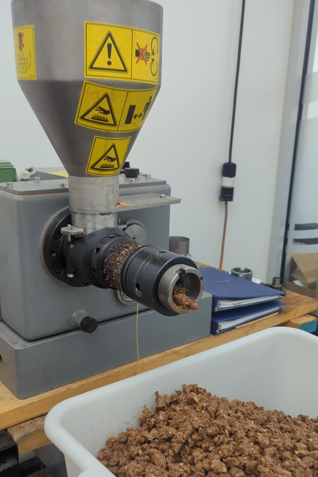 A machine used for processing or grinding material, with warning labels on it and brown processed material in a white container below.
