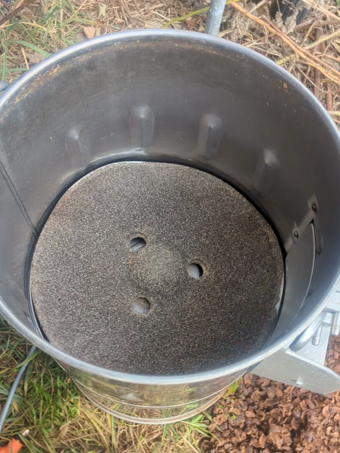 Empty stainless steel potato peeler with a textured surface inside, set outdoors on grass and rocks.