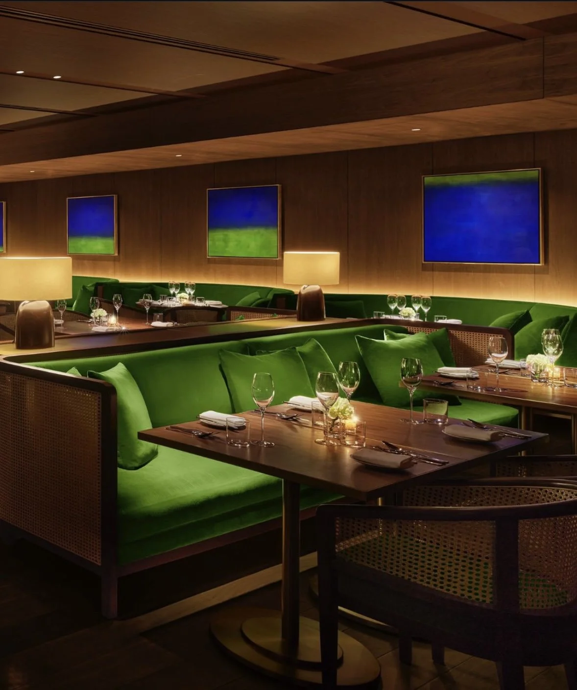 Elegant restaurant interior with green upholstered booths, wooden tables, and abstract landscape artwork on wooden-paneled walls, illuminated by warm lighting and table lamps.