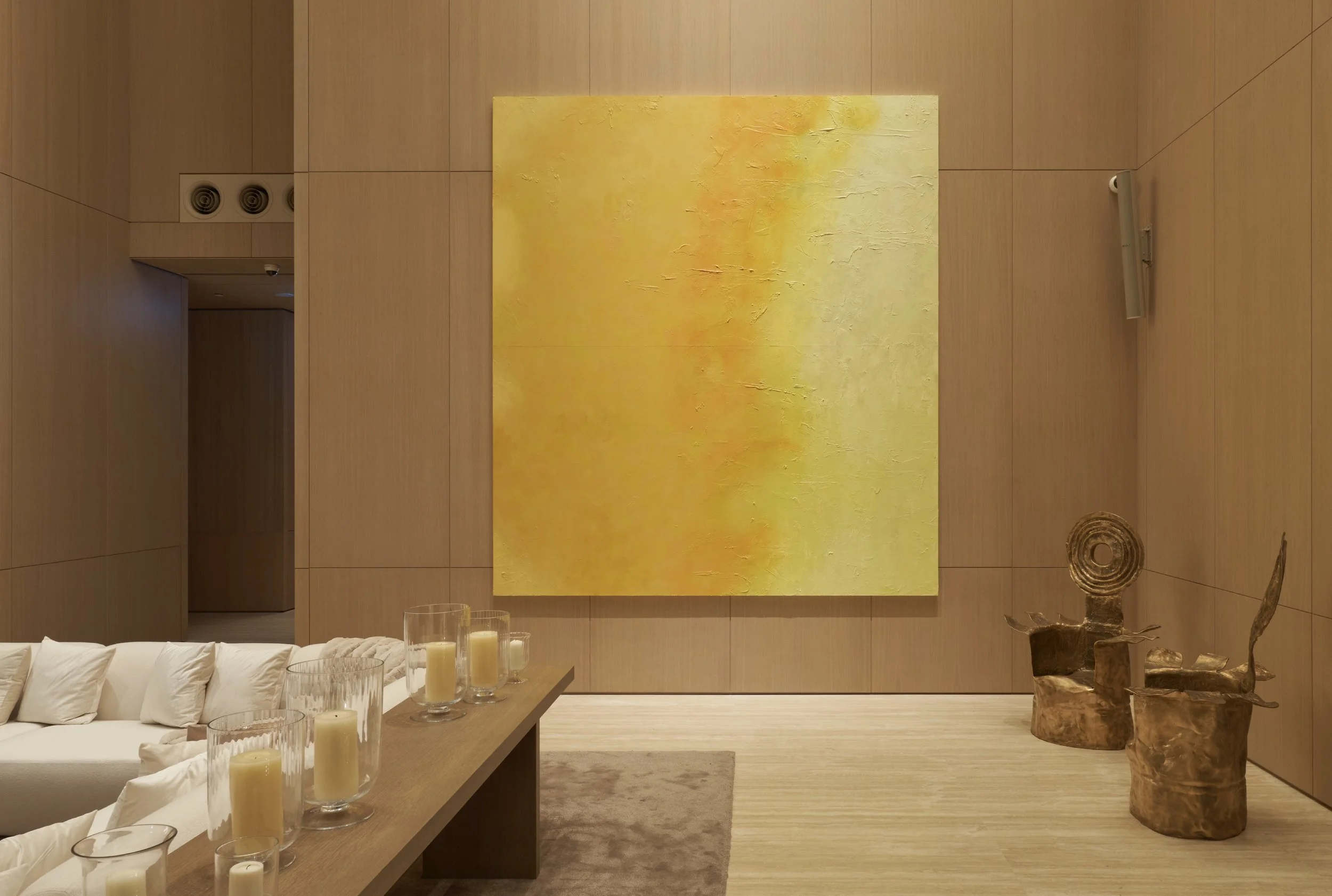 A modern interior with wooden walls, a large yellow abstract painting, a beige sofa with white pillows, a wooden table with multiple candles in glass holders, and two wooden sculptures shaped like chairs.